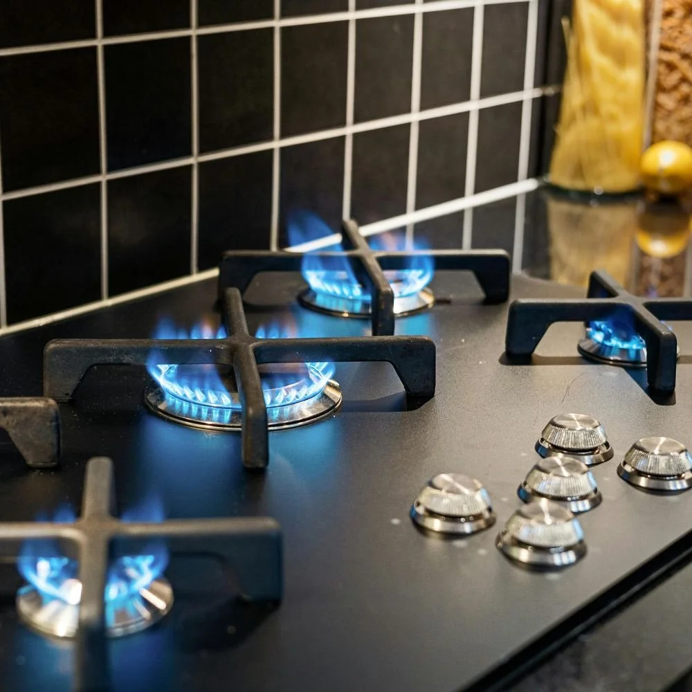 A modern gas stove with three lit burners and a black countertop, with black tiled backsplash and jars of pasta and grains in the background.