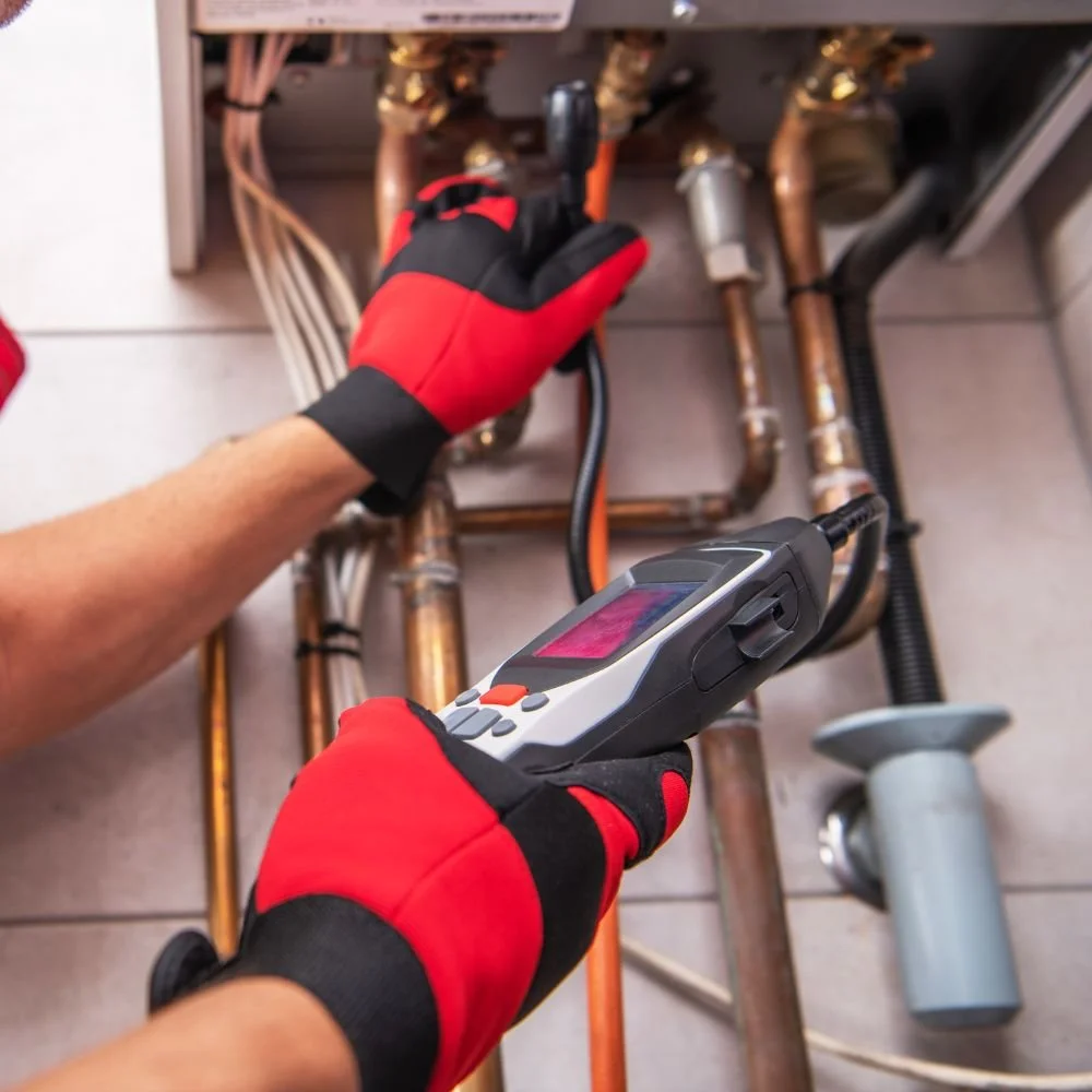 A technician wearing red and black gloves inspecting copper pipes beneath a sink with a handheld electronic testing device.