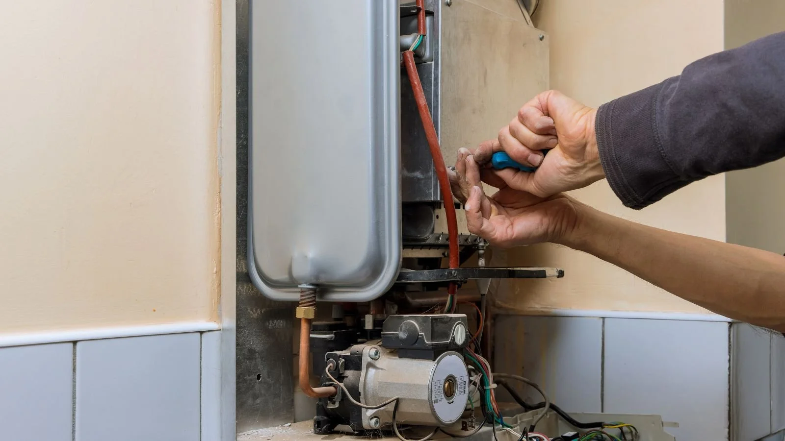 Person working on an electrical or plumbing appliance, using a screwdriver, with exposed wiring and mechanical components.