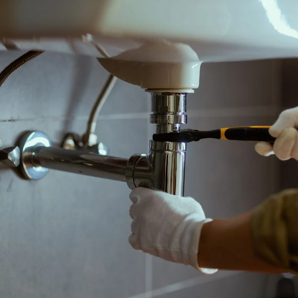 A person wearing white gloves is using a screwdriver to install or repair a chrome plumbing pipe underneath a sink.