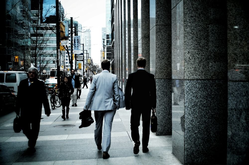Two men in suits walking on a city sidewalk, surrounded by office buildings and pedestrians.