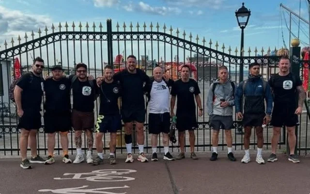 A group of ten men standing in front of a decorative gate at a sports venue, dressed in athletic clothing, with some wearing matching shirts, on a paved area under a partly cloudy sky.