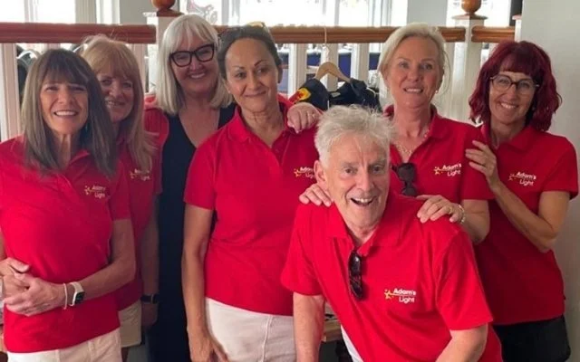 Group of six women and one elderly man smiling, wearing matching red shirts with a logo, indoors near a clothing rack.