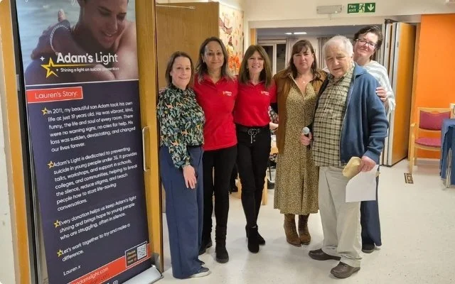 Group of six people standing in a hallway near a poster board, with one person holding a microphone and paper, during a community event.