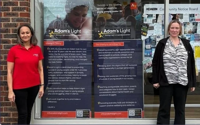 Two women stand in front of a large informational display about Adam's Light, a suicide prevention organization. The woman on the left wears a red jacket, and the woman on the right wears a black blazer and a blouse with a pattern. The display contains text about the organization's mission and Story, with a background showing a person writing on a translucent surface.