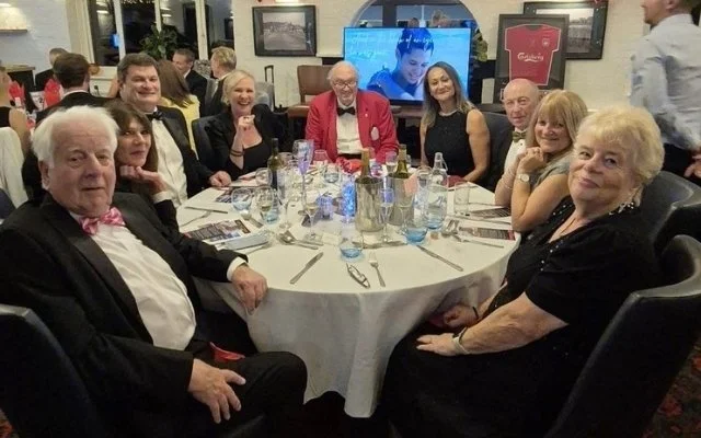 A large group of people sitting around a round dining table at a restaurant with a TV screen in the background.