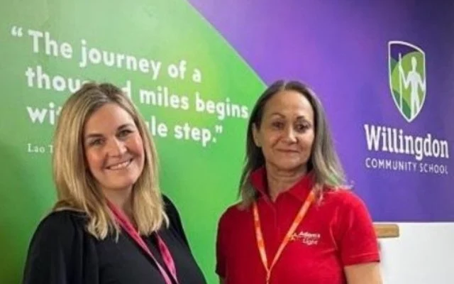 Two women standing in front of a green and purple wall with a quote and the Willingdon Community School logo. The quote says, 'The journey of a thousand miles begins with a single step.'