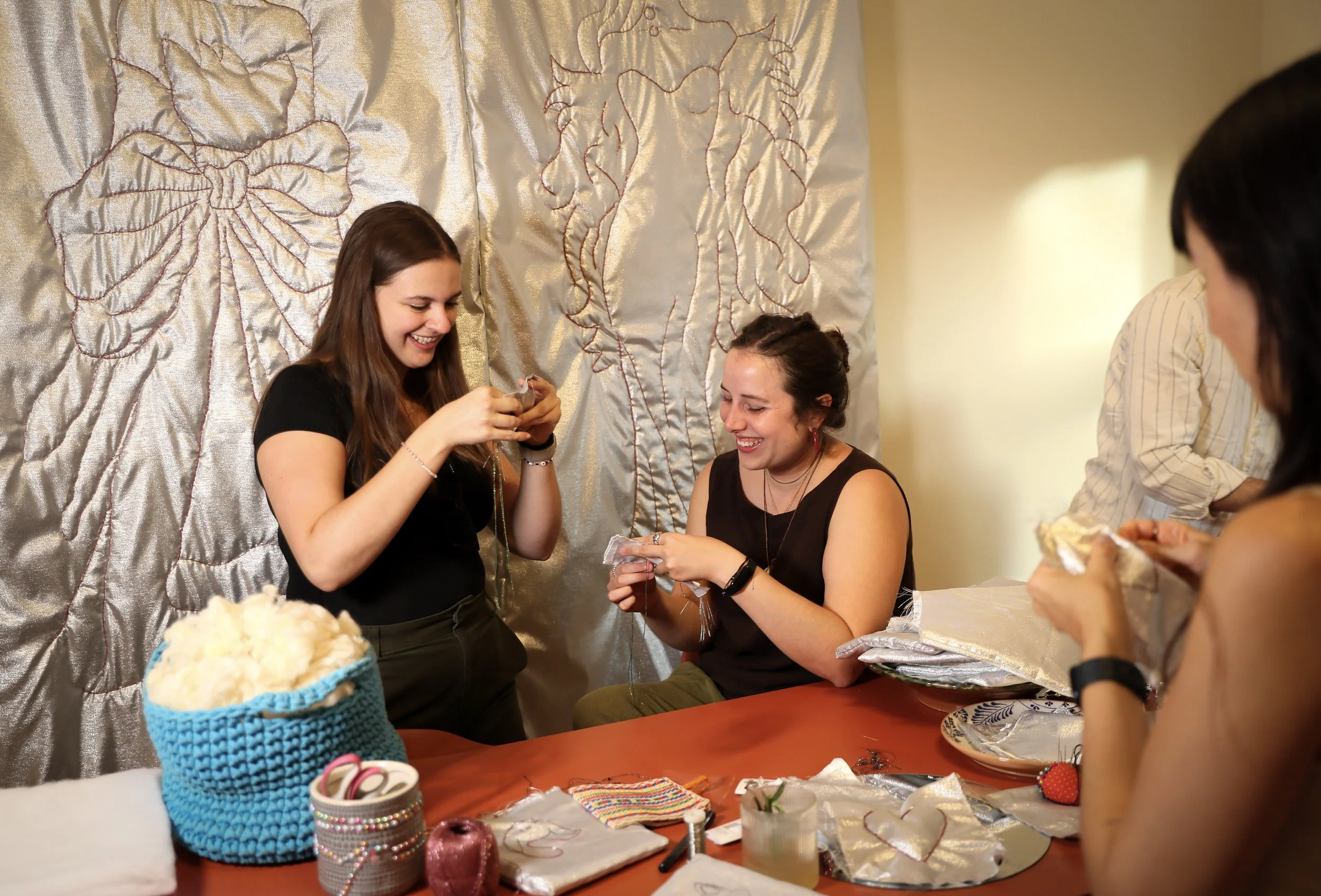 Vier Frauen sind in einem Raum, der mit einer großen silbernen Decke mit Goldstickerei dekoriert ist, rund um einen Tisch versammelt. Sie scheinen Geschenke zu öffnen und lachen. Der Tisch ist mit Bändern, Stoffen und einem Korb mit Häkelarbeiten bedeckt.