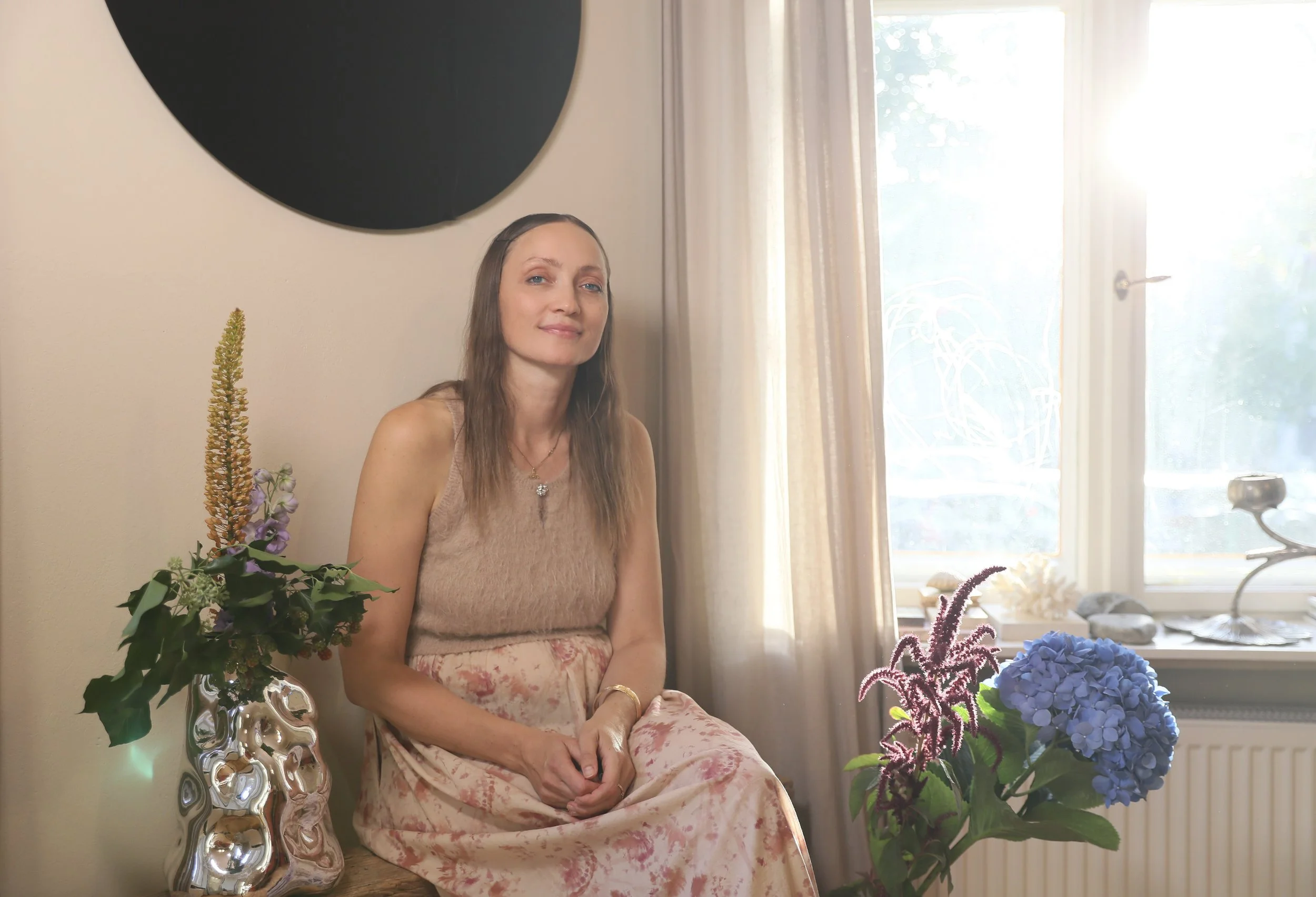 Eine Frau sitzt in einem Raum neben einem Regal mit blühenden Blumen, Sonnenlicht scheint durch ein Fenster im Hintergrund.