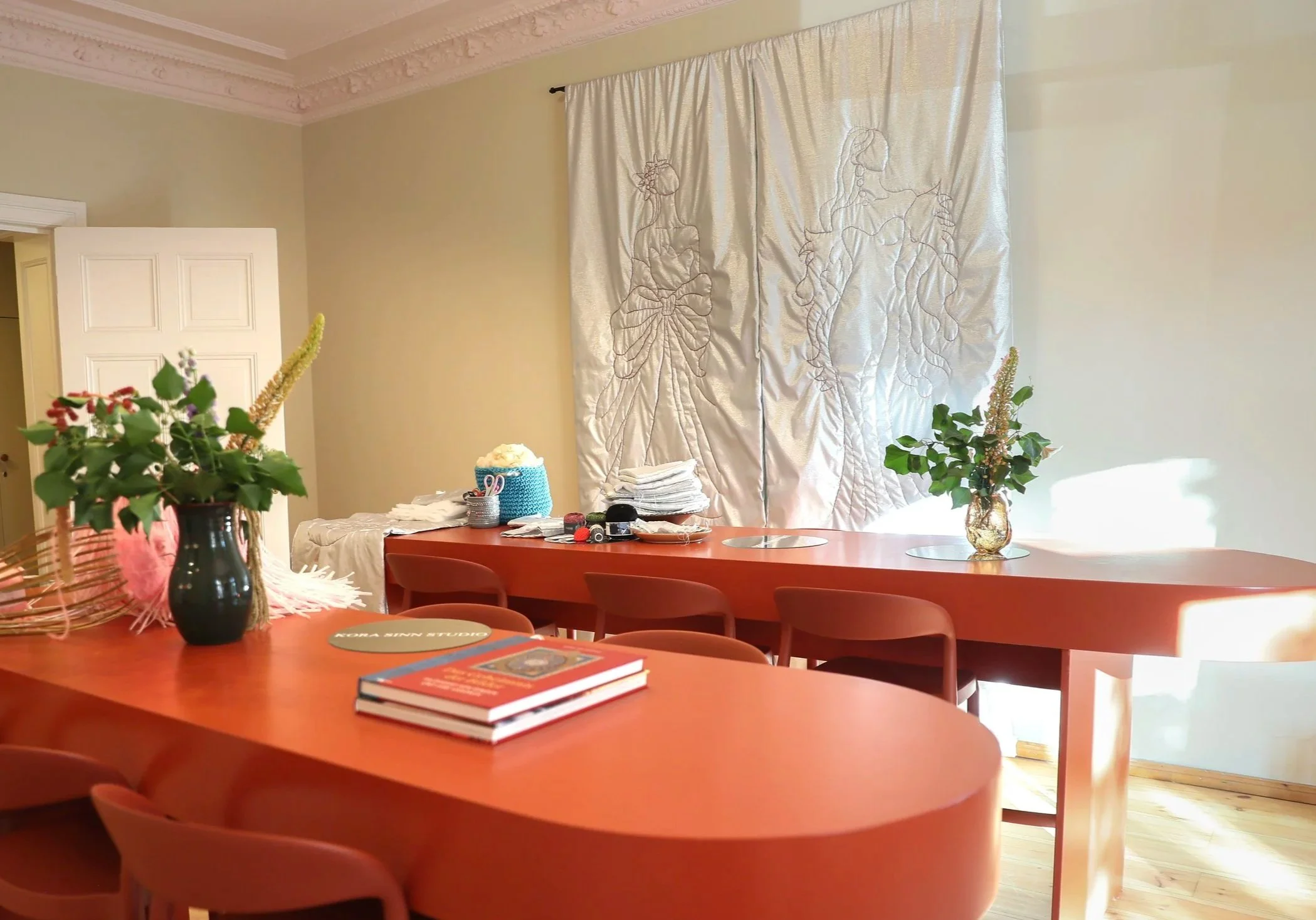 Ein Esszimmer mit einem großen, ovalen, orangefarbenen Tisch und mehreren roten Stühlen. Es gibt mehrere Pflanzen auf dem Tisch und auf einer Seite des Raumes, sowie Bücher und Kosmetika. Die Wand hat eine große, seidene Gardine mit Stickerei von zwei Zigeunertänzinnen.