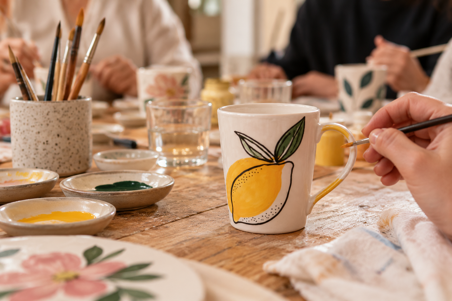 Menschen beim Keramikbemalen an einem Holztisch, eine Person bemalt eine weiße Tasse mit einer Zitronendekoration, umgeben von Malfarben, Pinseln, Wasserbechern und Geschirr mit Blumenmuster.