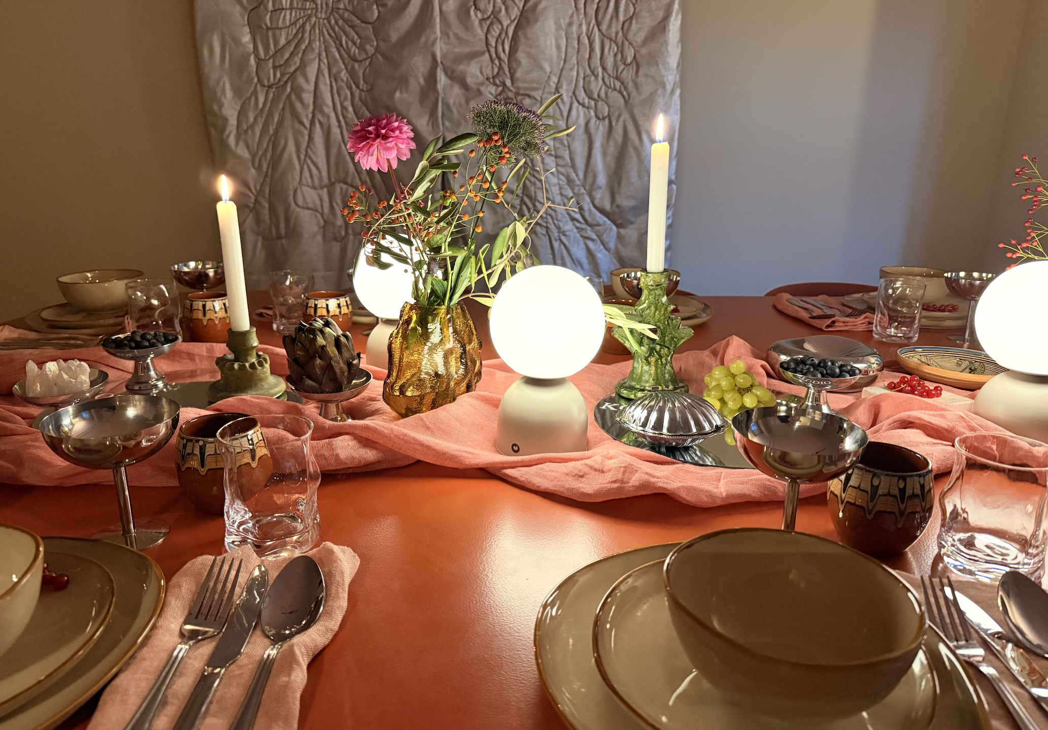 Ein festlich gedeckter Esstisch mit Kerzen, Blumenstrauß und verschiedenen Geschirr- und Gläserwaren, auf einem orangefarbenen Tuch. Im Hintergrund eine gepolsterte Rückenlehne.