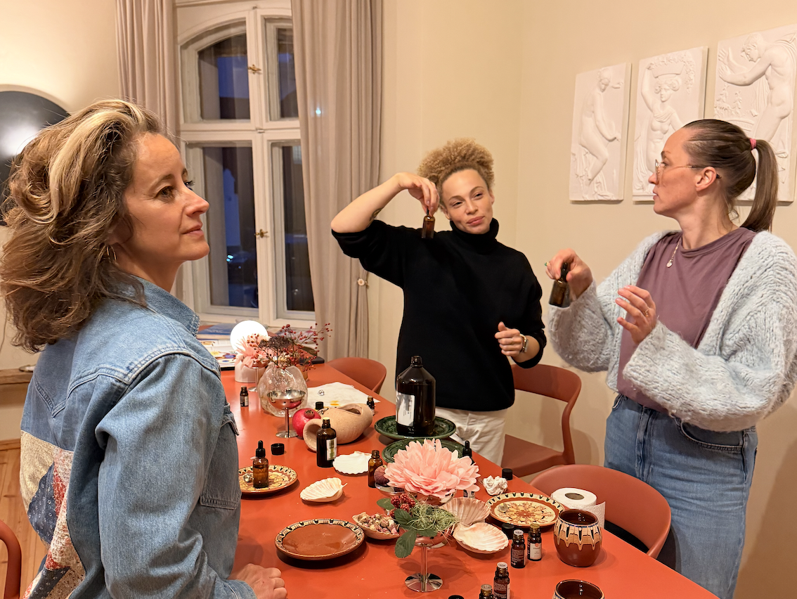 Drei Frauen in einem Wohnzimmer, die sich um einen Esstisch versammelt haben. Eine Frau links trägt eine Jeansjacke, die andere in der Mitte hält eine kleine Flasche hoch, und die rechte Frau trägt eine gestrickte Jacke und eine T-Shirt. Der Tisch ist mit Dekorationen, Flaschen, kleinen Gegenständen und Küchenware geschmückt.