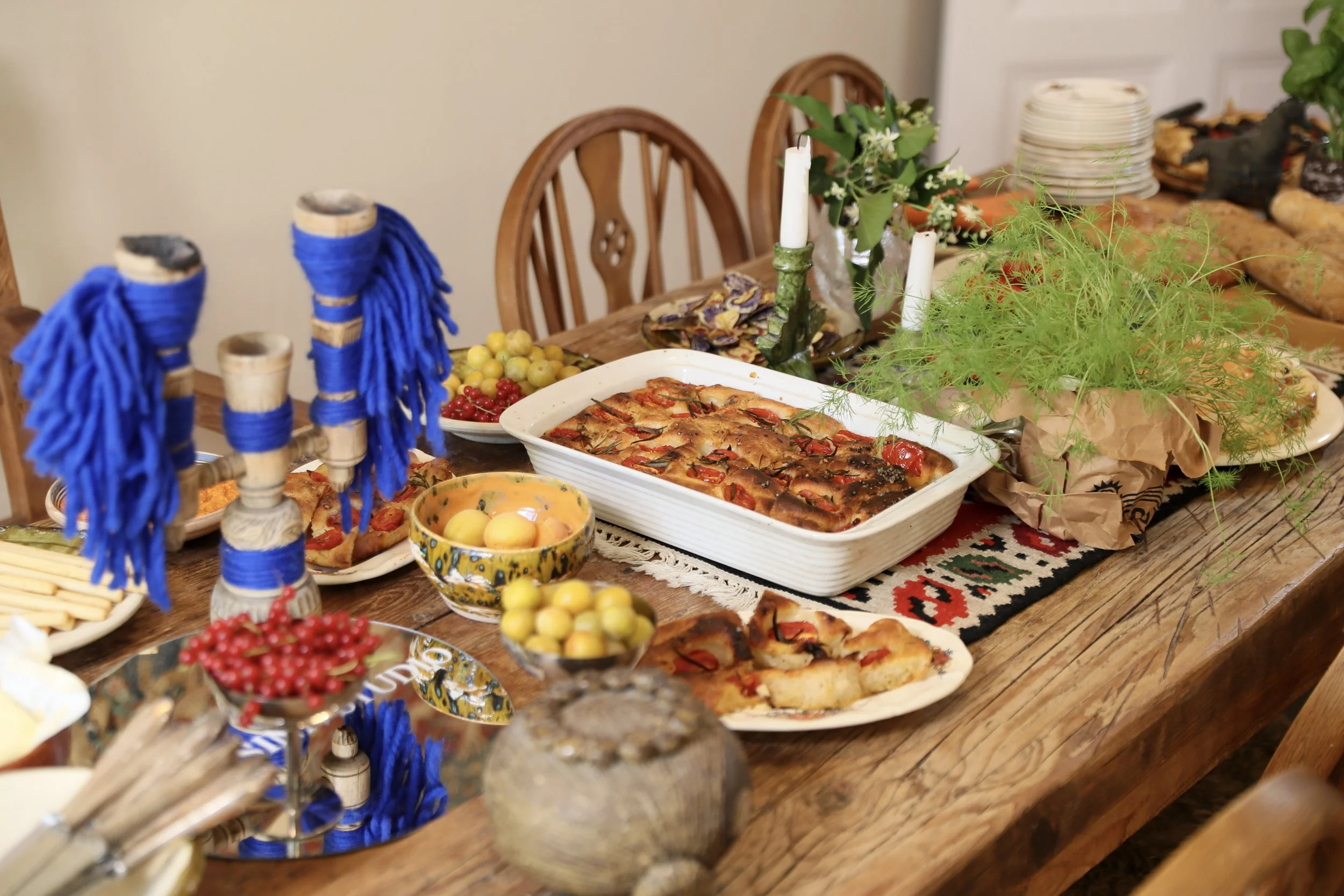 Ein festlich gedeckter Tisch mit verschiedenen Speisen und Dekorationen, inklusive Ofenkuchen, Obst, Kerzen und Blumen, auf einem traditionellen Teppich.