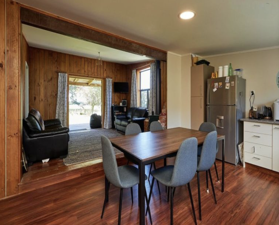 Open-concept kitchen and living room with wooden walls and floors, dining table with six chairs, refrigerator, kitchen counter with appliances, and seating area with sofas near large windows.