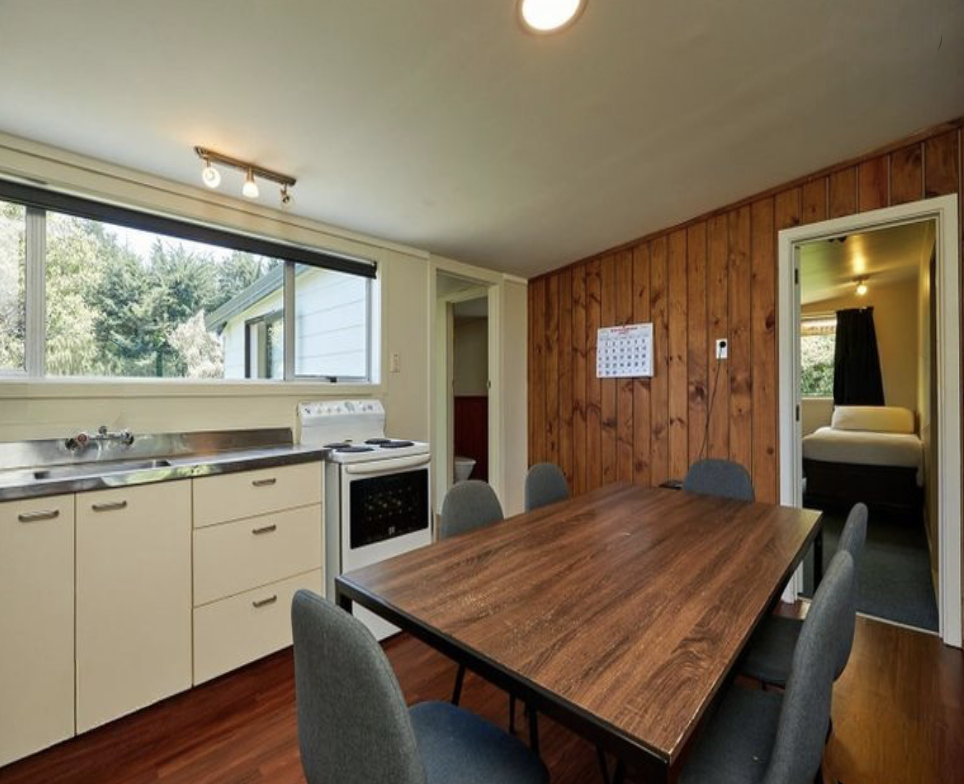 Kitchen and dining area with a wooden table, six gray chairs, a window with a view of greenery, a white stove, and a wooden paneled wall. A doorway leads to a bedroom with a bed and dark curtain.