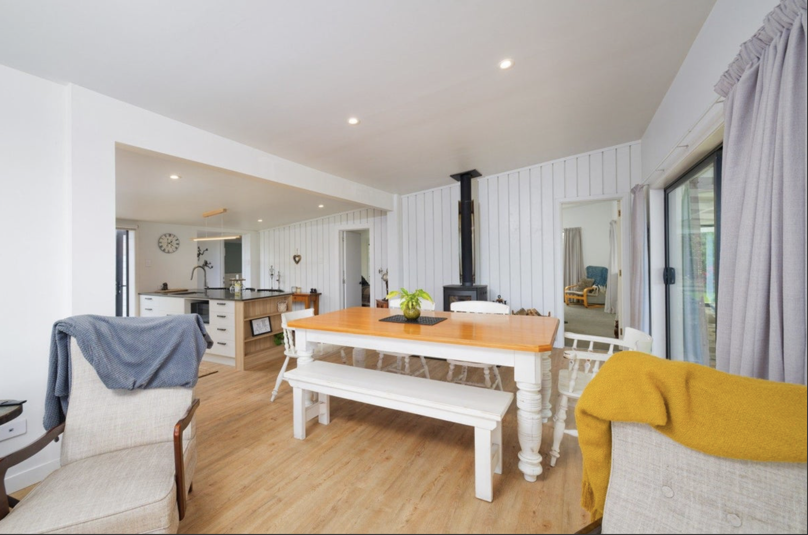 Open-plan living and dining area with white walls, wooden flooring, and large sliding glass doors. It features a wooden dining table with benches and chairs, a cozy armchair with a gray blanket, and a corner with a wood stove. The space is bright and