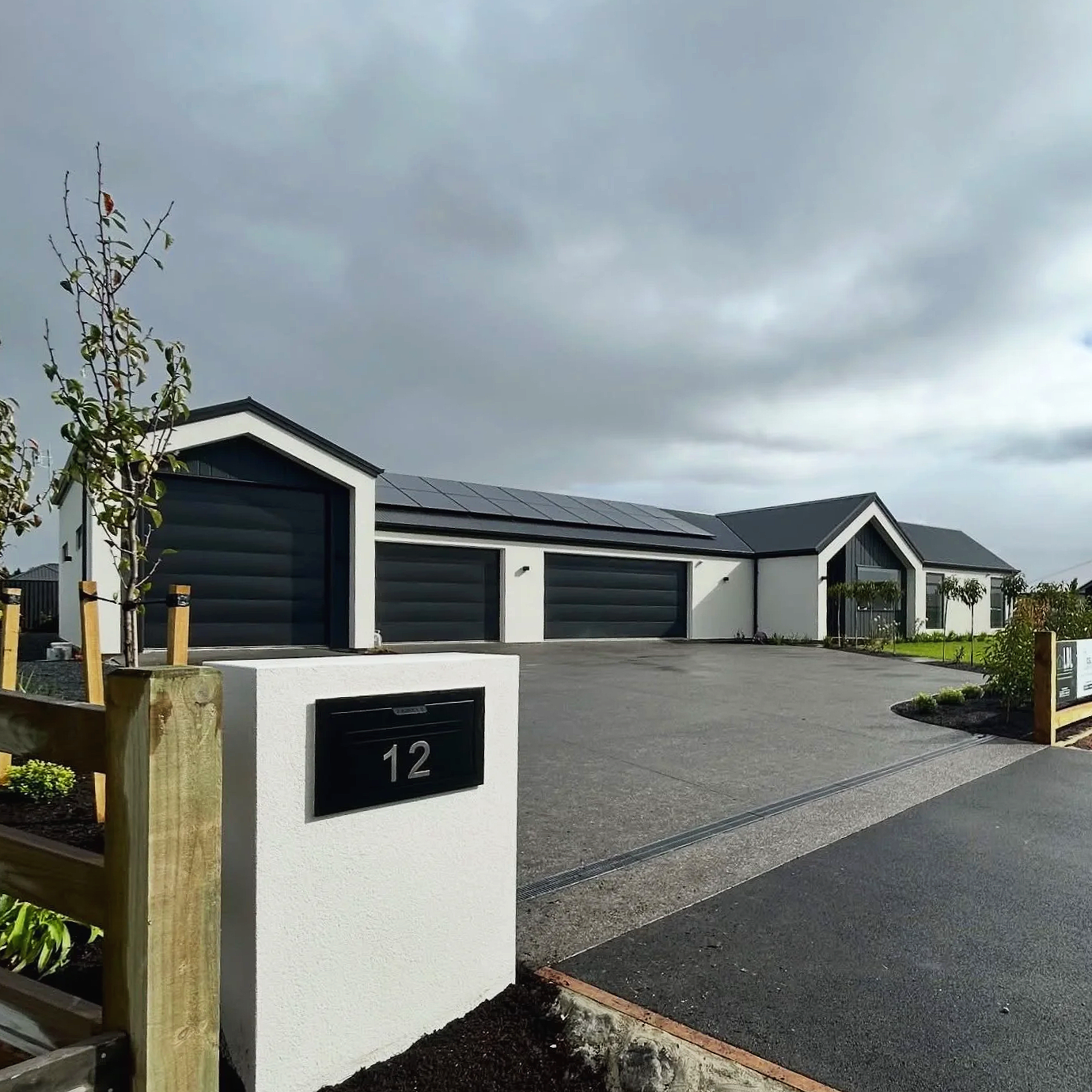 Modern residential house with white walls, black garages, and solar panels on the roof, with a driveway and landscaped front yard under cloudy sky.