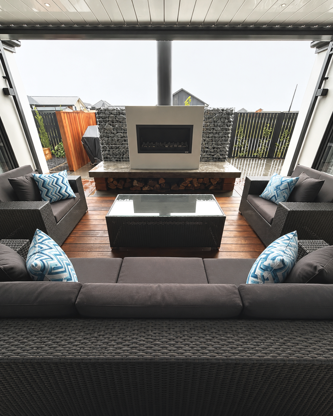 Outdoor living space with wicker sofas and armchairs with blue patterned pillows, a glass-topped coffee table, and a modern fireplace on a wooden deck. Fenced backyard with a barbecue grill and neighboring houses.
