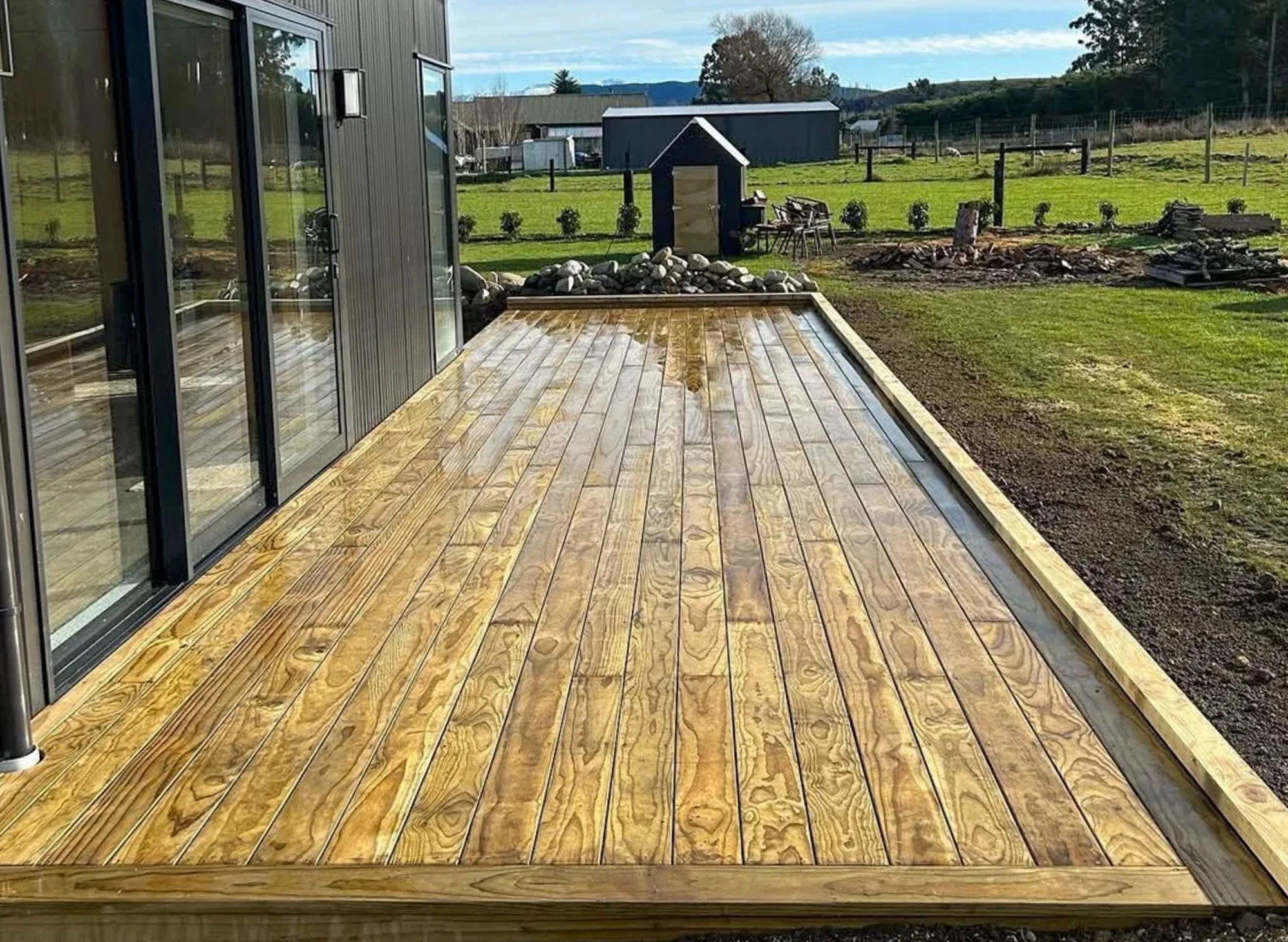 Wooden deck outside a house with sliding glass doors, surrounded by a grassy yard with small plants and a black shed in the background.