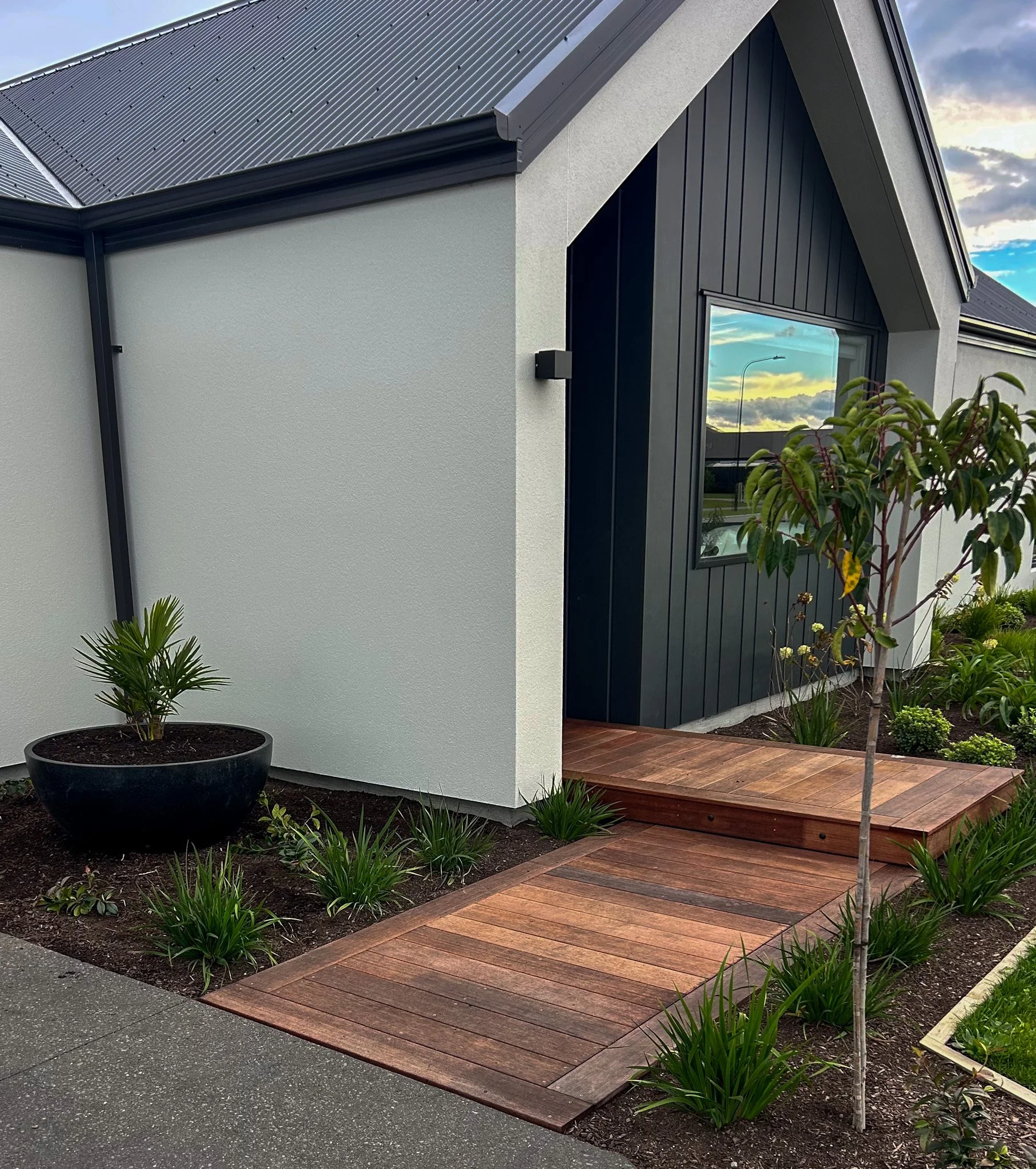 A modern house exterior with white and black walls, a wooden porch, and landscaped garden with greenery, small trees, and shrubs.