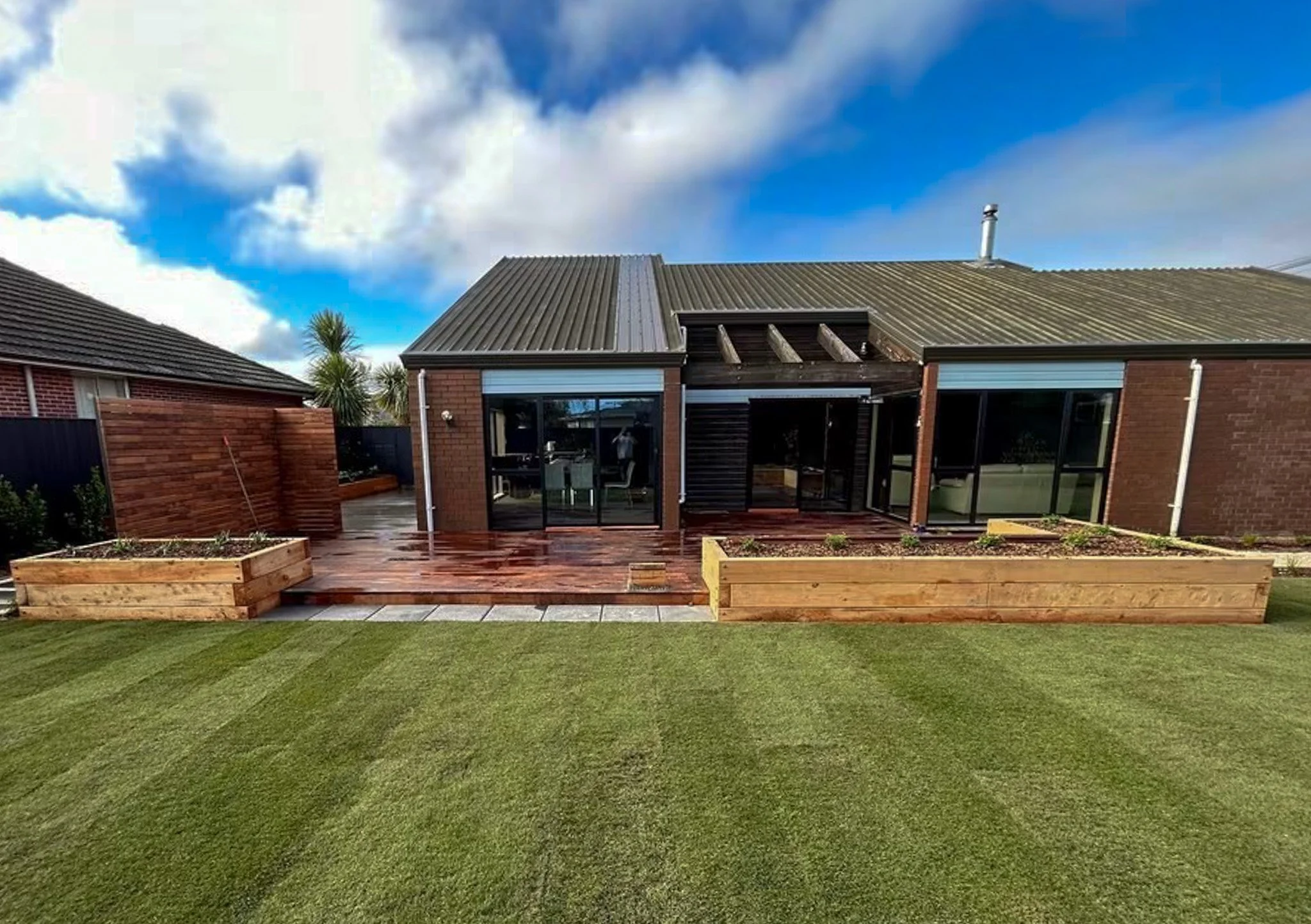 Backyard with a wooden deck, green grass, two raised garden beds, brick house with glass doors, cloudy sky.