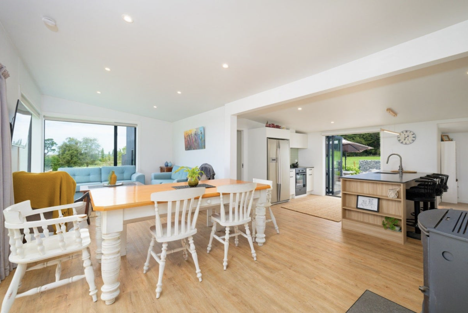 Open-concept kitchen and living room with white walls, wooden flooring, large windows, and sliding glass doors leading to outdoor greenery.