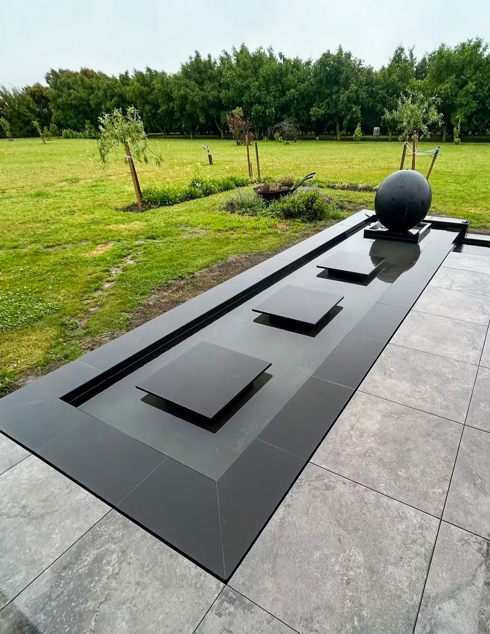 A modern sculpture of a black sphere on a rectangular base, located outdoors on a paved area, with a grassy field and trees in the background.