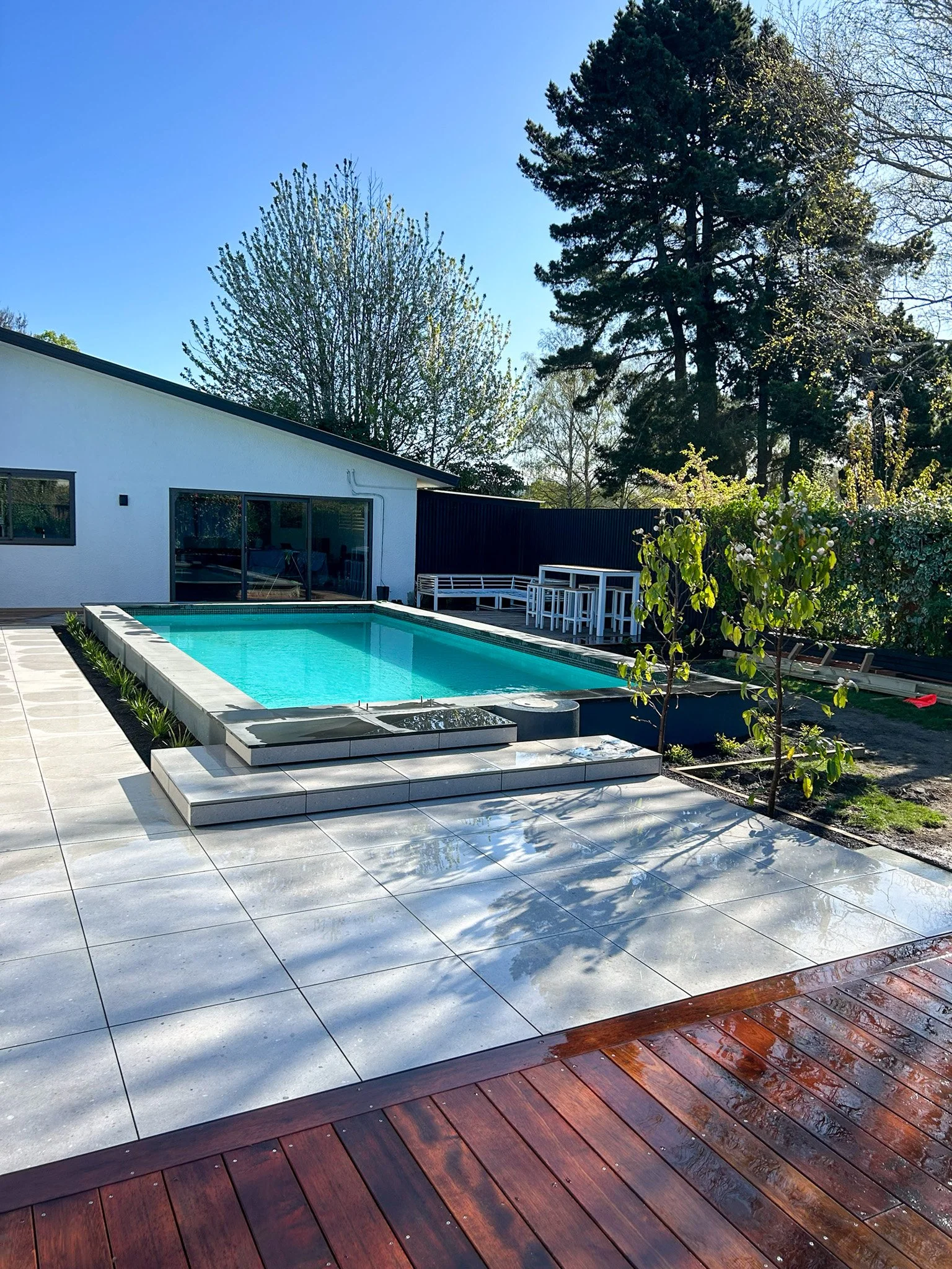 Backyard with a swimming pool, white and dark outdoor furniture, trees, and a clear blue sky.