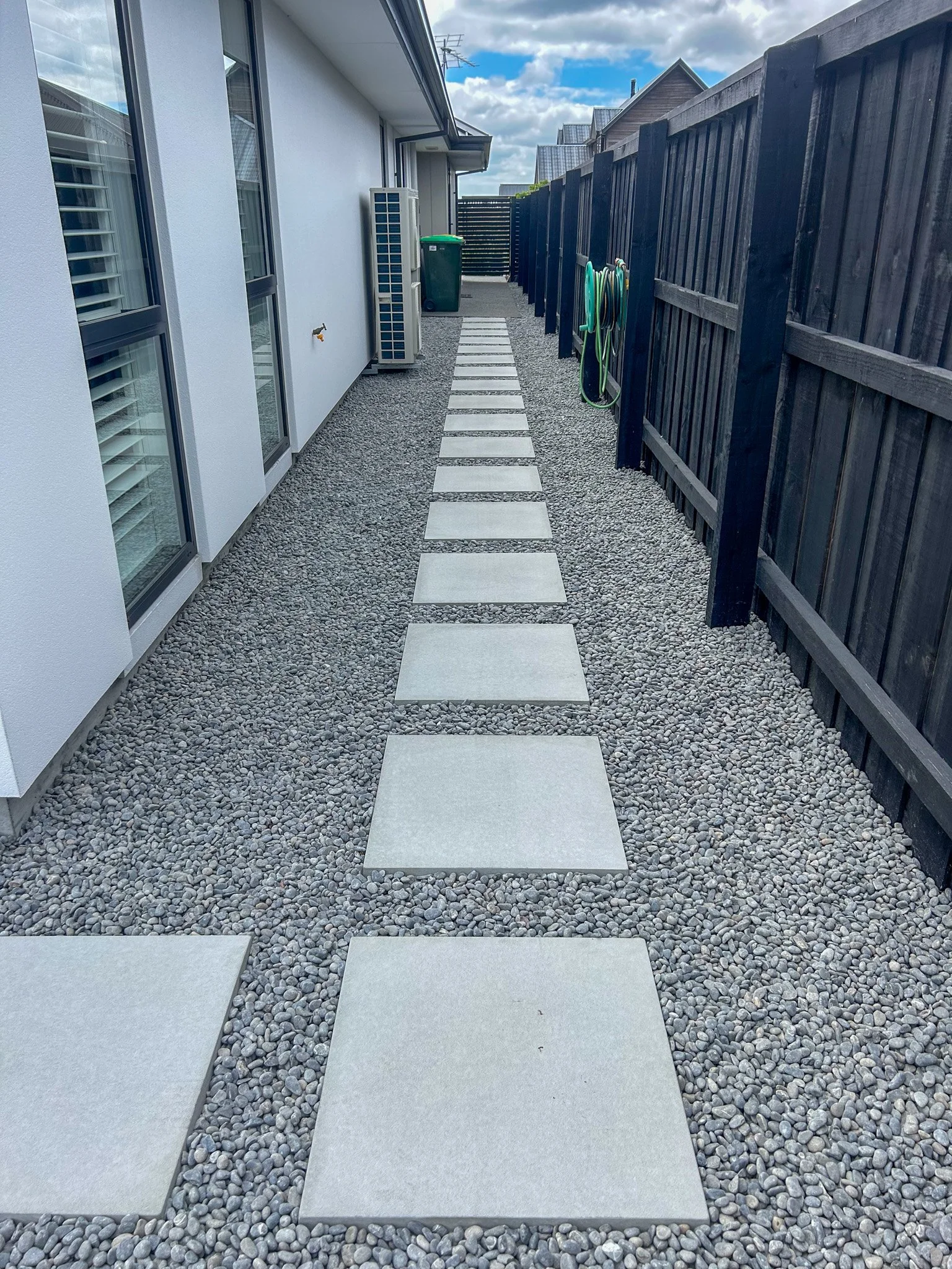 Backyard pathway with square concrete pavers on gravel, bordered by a black wooden fence on the right and a white house wall with windows and an air conditioning unit on the left. Garden hoses are hanging on the fence, and a green trash bin is at the