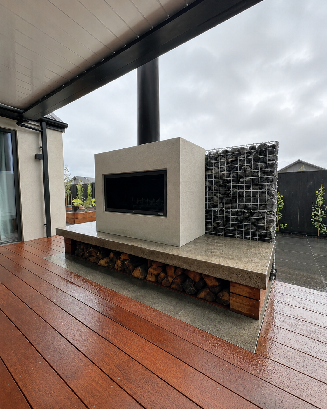An outdoor patio with rain-wet wooden flooring, modern fireplace with a black glass front, a decorative gabion filled with rocks behind it, and stacks of firewood underneath the fireplace. There's a black railing and some potted plants in the background.
