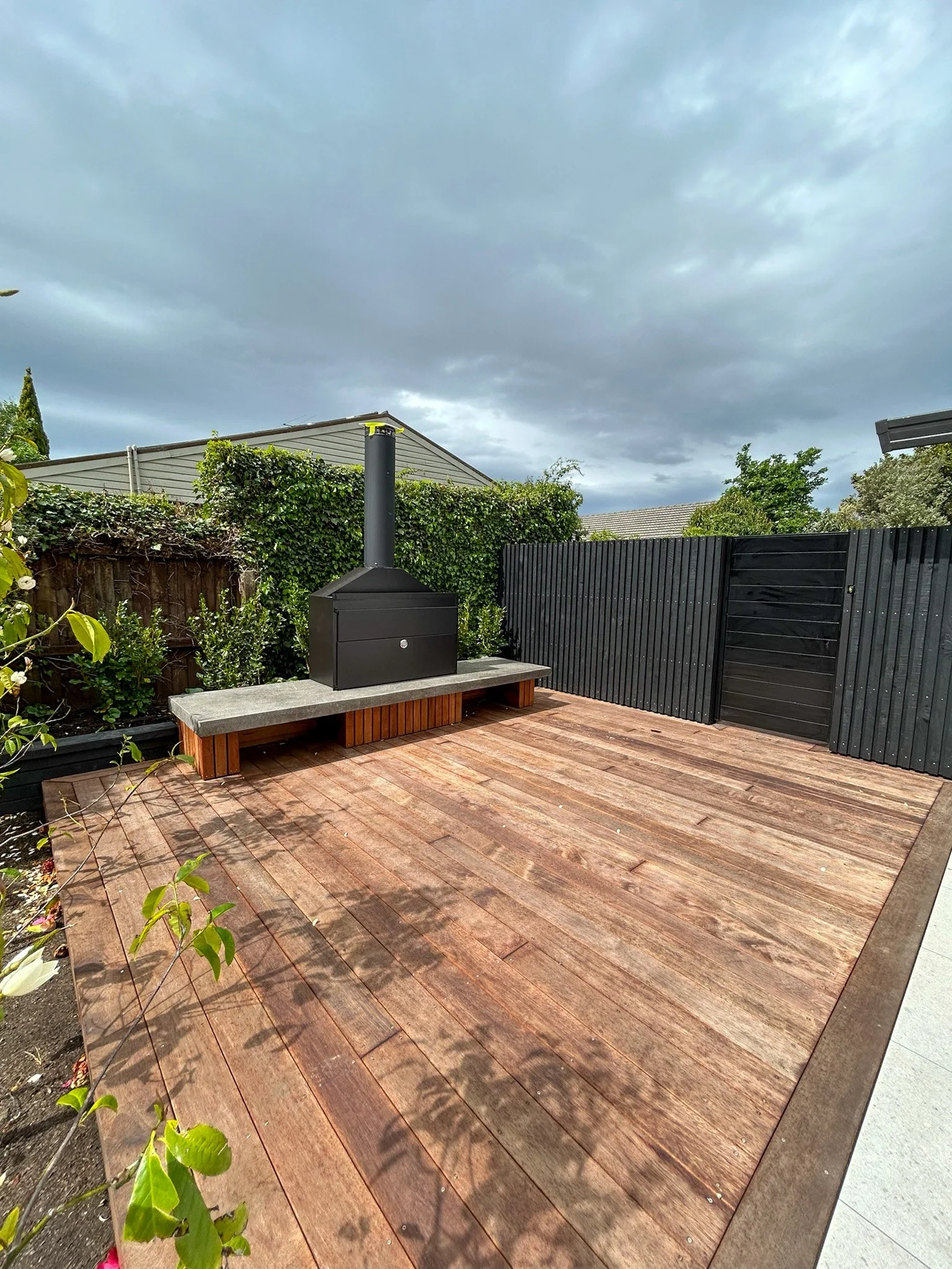 A backyard patio area with wooden decking, a black outdoor stove, and a black fence, with greenery and bushes along one side and cloudy skies overhead.