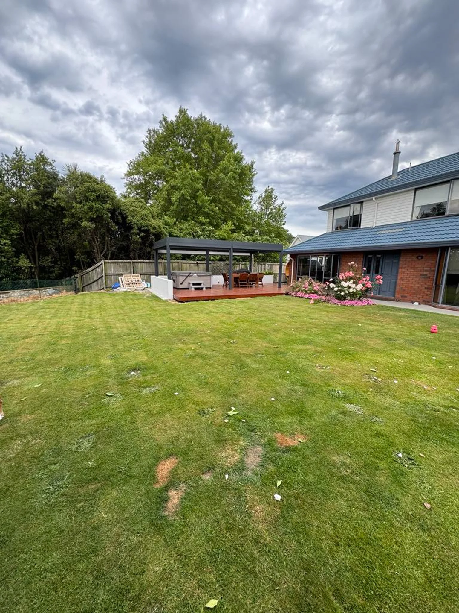 A backyard with a green lawn, a covered patio area with outdoor furniture, a flower bed with pink and white flowers, and a two-story house with white siding and a blue roof. There are trees and cloudy skies in the background.