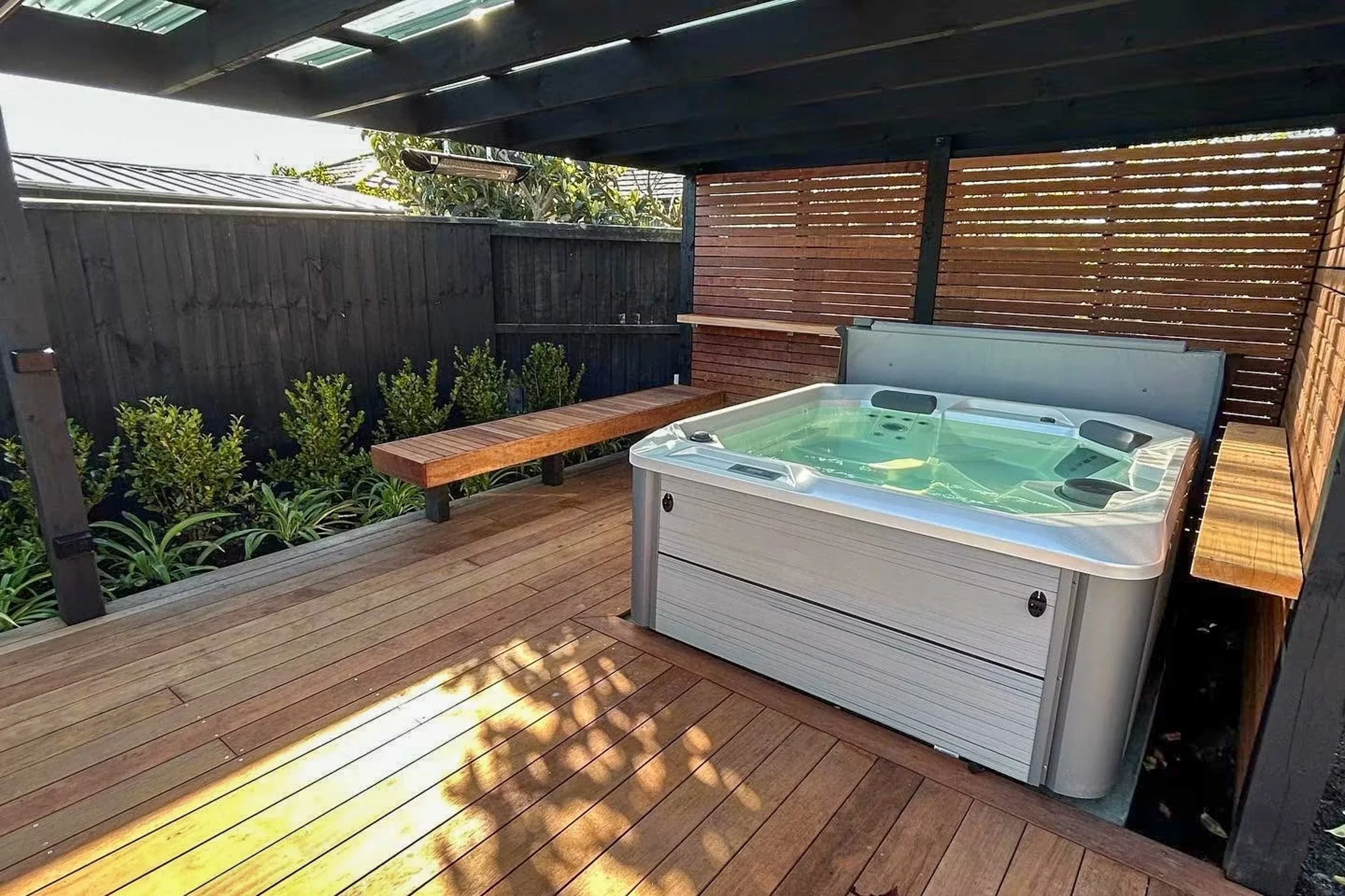 A outdoor wooden deck with a hot tub filled with water, surrounded by a black and wood privacy fence, greenery, and a roof overhead, with sunlight casting shadows on the deck.