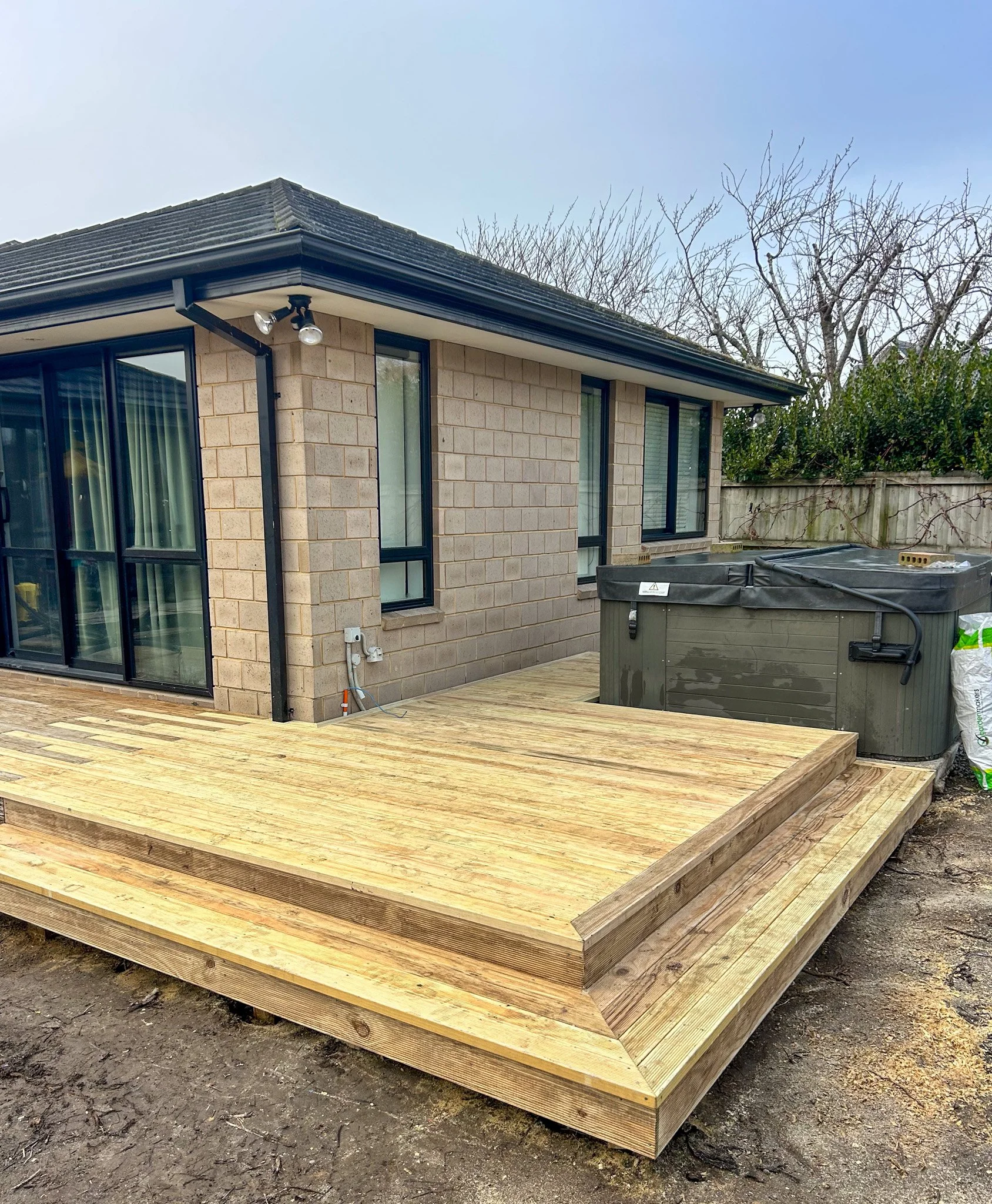 Backyard deck under construction with new wooden flooring, house with large windows and brick exterior, and a backdrop of leafless trees and a clear blue sky.