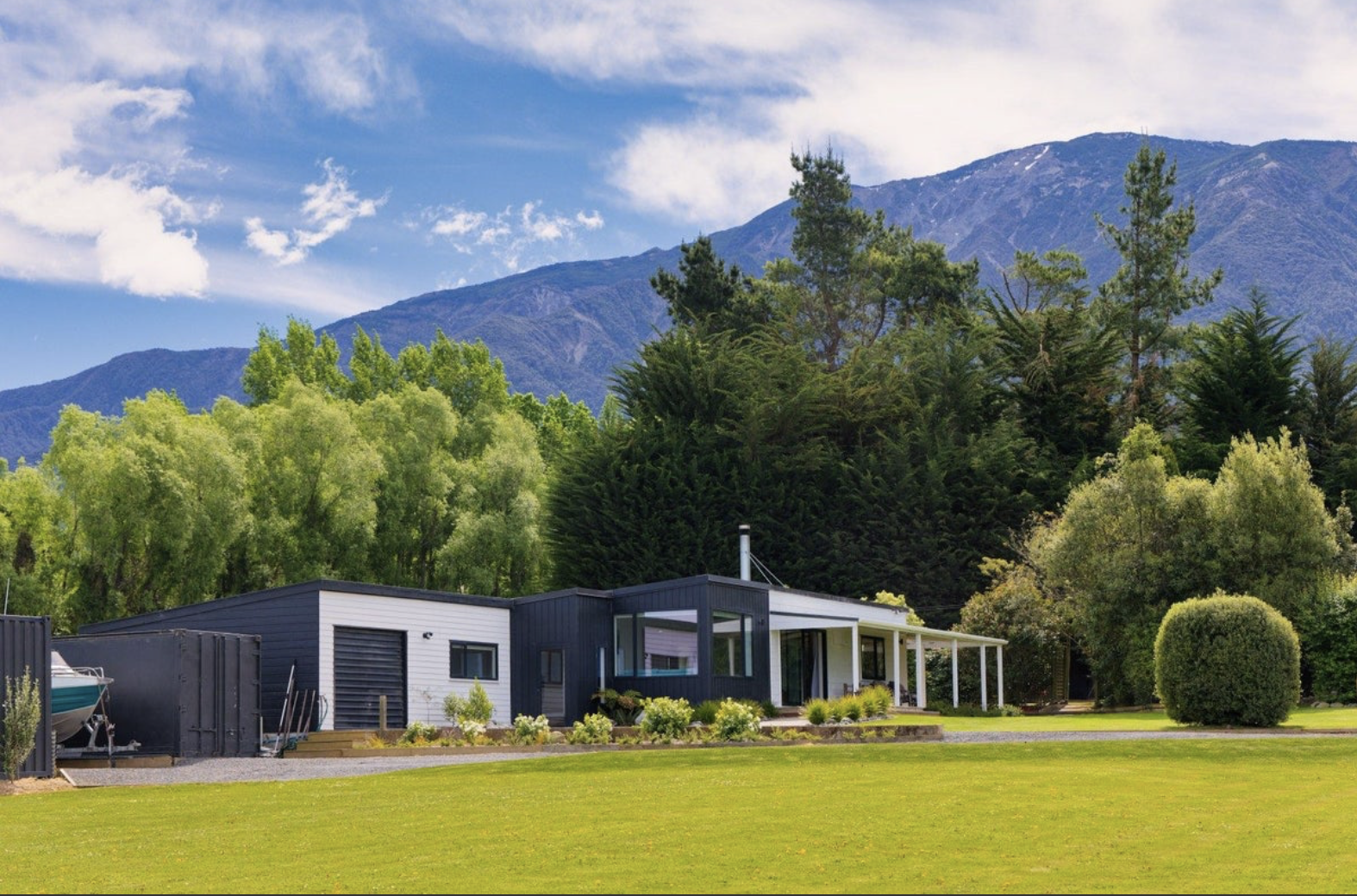 Modern house with black and white exterior in front of a lush green lawn, surrounded by trees and mountains in the background under a partly cloudy sky.