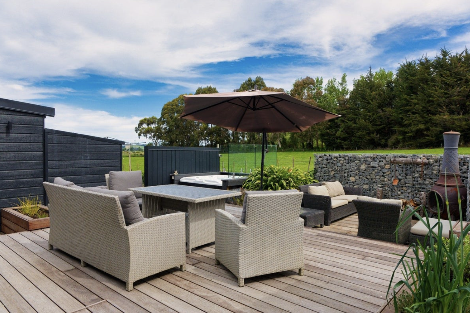 Outdoor patio with wicker and fabric furniture, a large umbrella, a hot tub, and a wood burning stove, surrounded by a black fence and lush green trees under a partly cloudy sky.