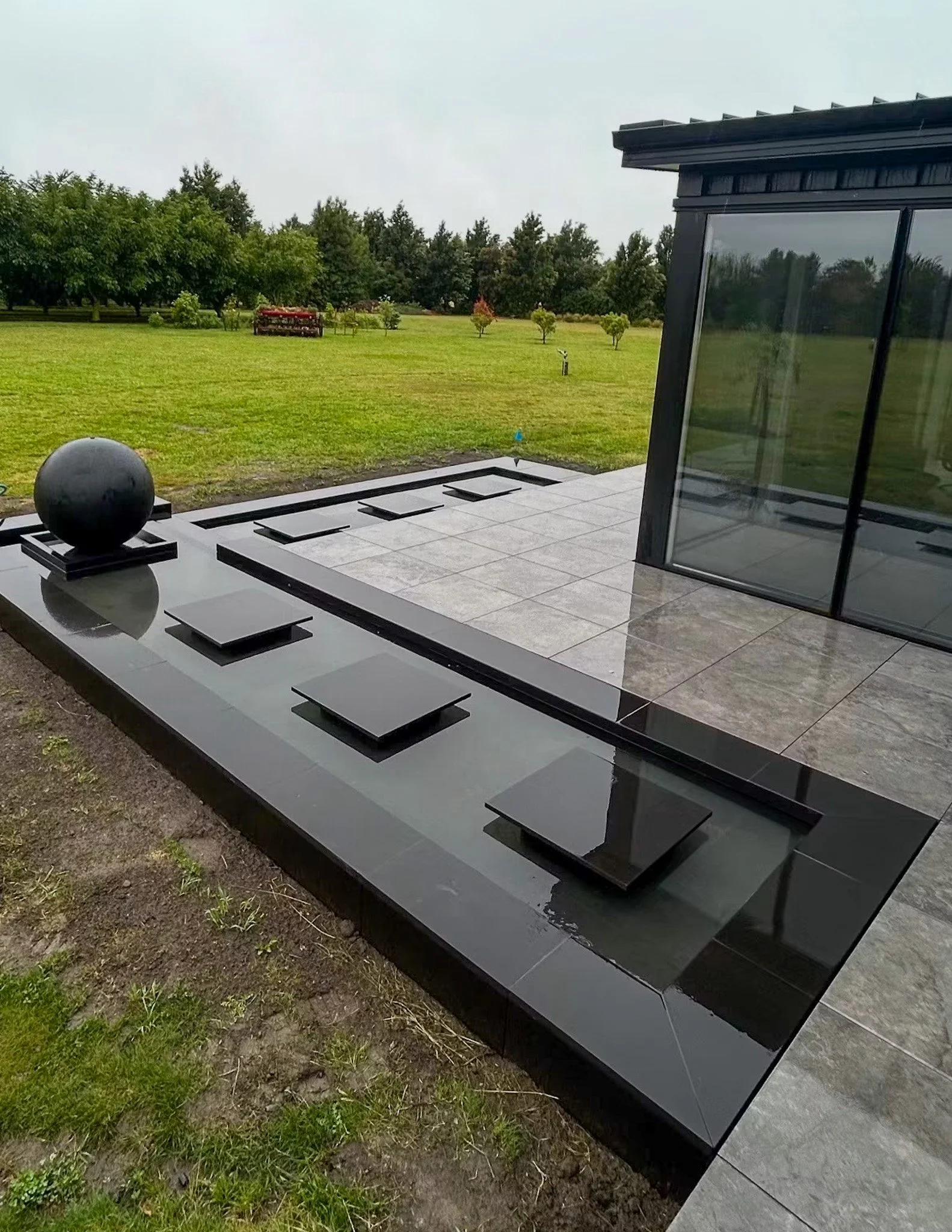 Modern black house exterior with a large reflective window, a spherical sculpture on a black slab, and a patio area with gray tiles. Green lawn with small trees and a red garden bed in the background.
