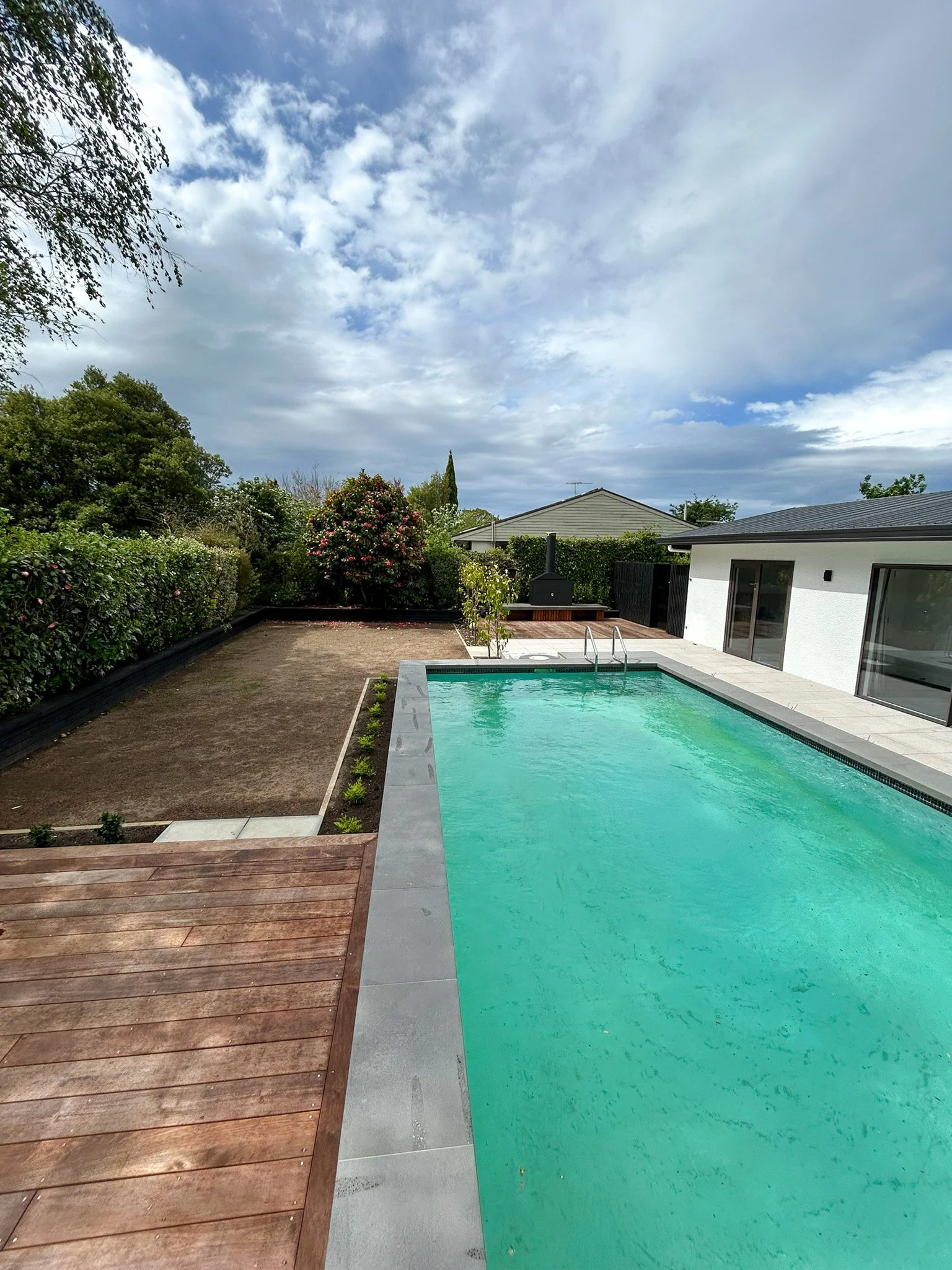 A backyard with a swimming pool, wooden deck, plants, and a white modern house with large glass doors, under a partly cloudy sky.