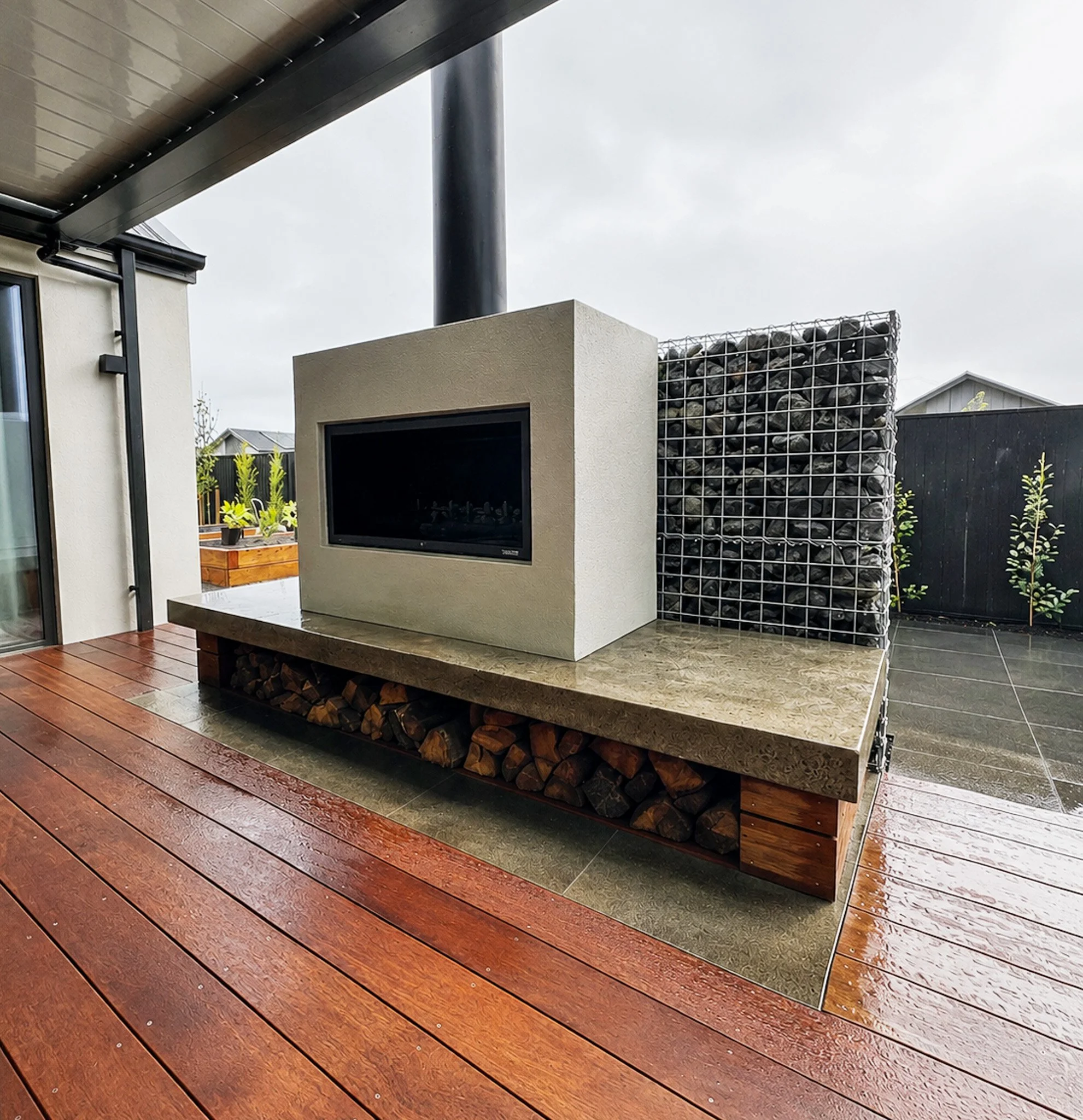 Close-up of an outdoor modern patio area with polished wooden flooring, a beige concrete fireplace with wood storage underneath, and a storage cage filled with rocks, with a house and cloudy sky in the background.
