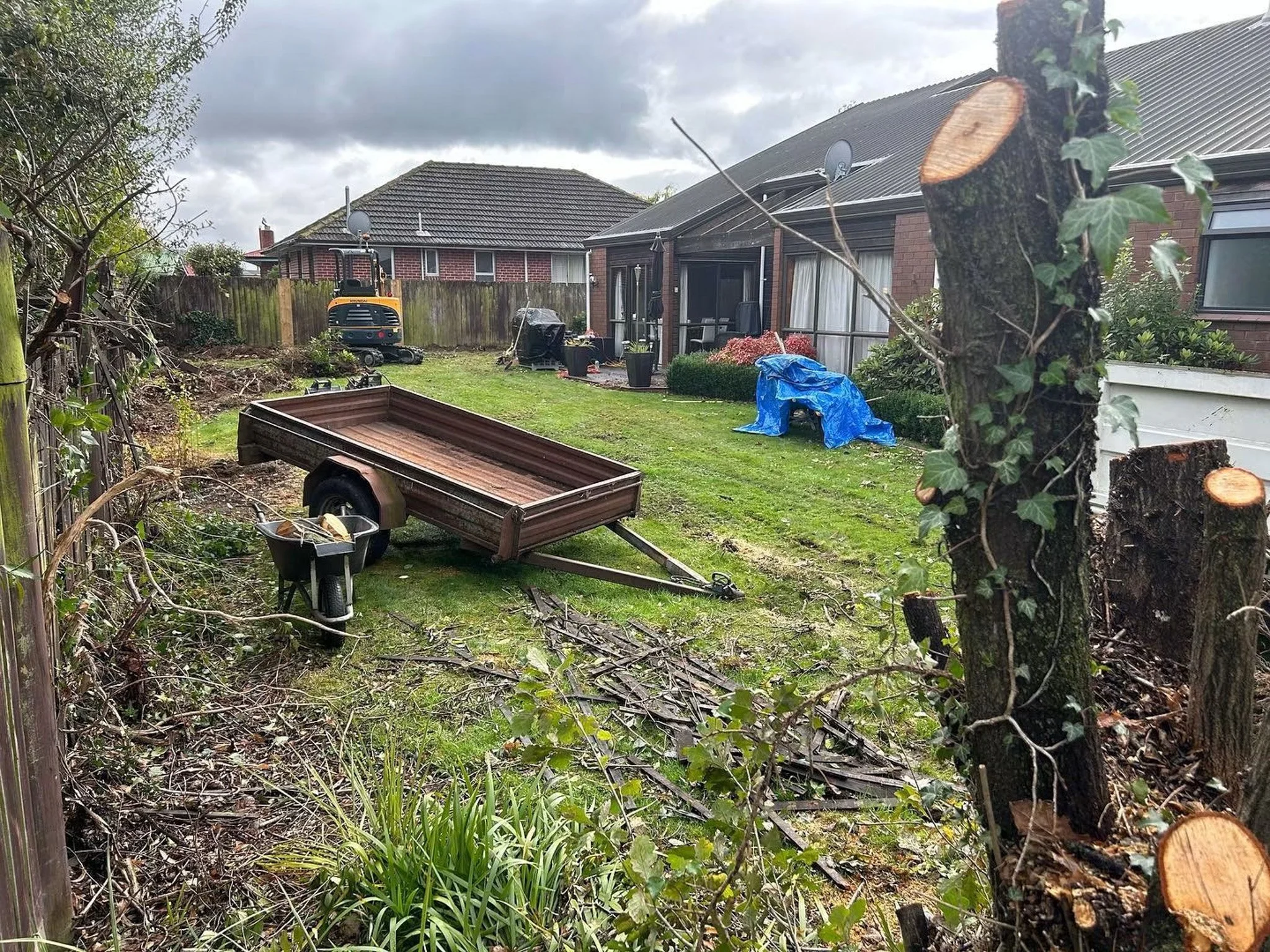View of a backyard with a grassy lawn, a broken down trailer, and various gardening tools. A tree trunk with ivy is in the foreground on the right, and a small plant is near the bottom left. Multiple houses with brick exteriors and tiled roofs are vi