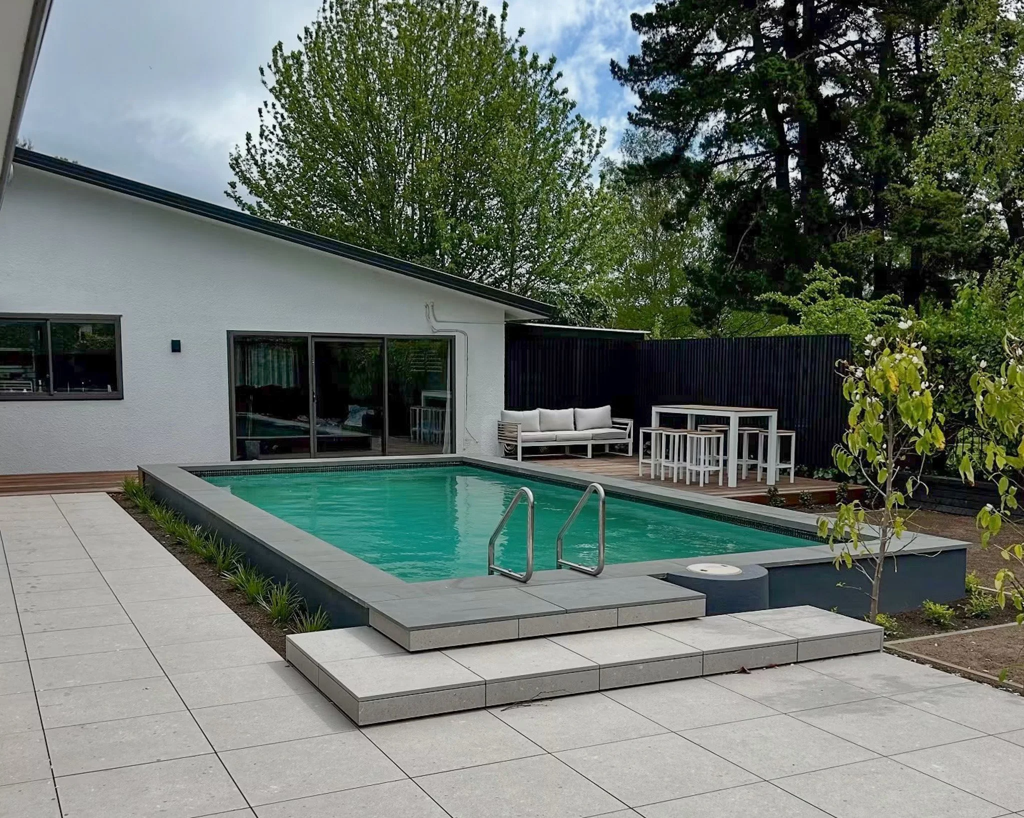 A modern backyard patio with a rectangular swimming pool, white outdoor furniture, a sliding glass door, and a white house wall. Tall trees and a partly cloudy sky are visible in the background.