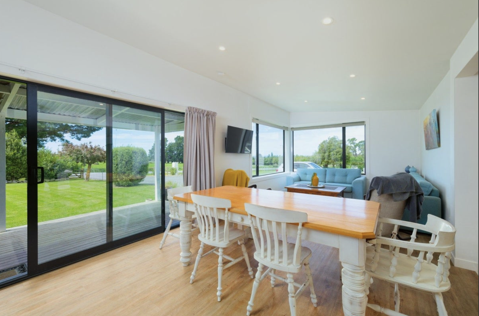 Open living and dining room with large sliding glass doors overlooking a green yard. The room features a wooden dining table with white chairs, a light blue sofa, a small coffee table, a wall-mounted TV, and a window with curtains.