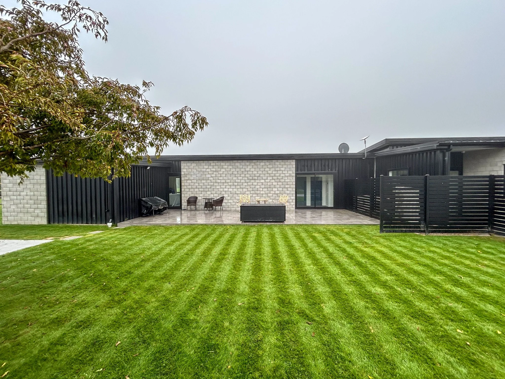 Modern house with black and white exterior, patio furniture, black privacy fence, tree on left, well-maintained green lawn, overcast sky.