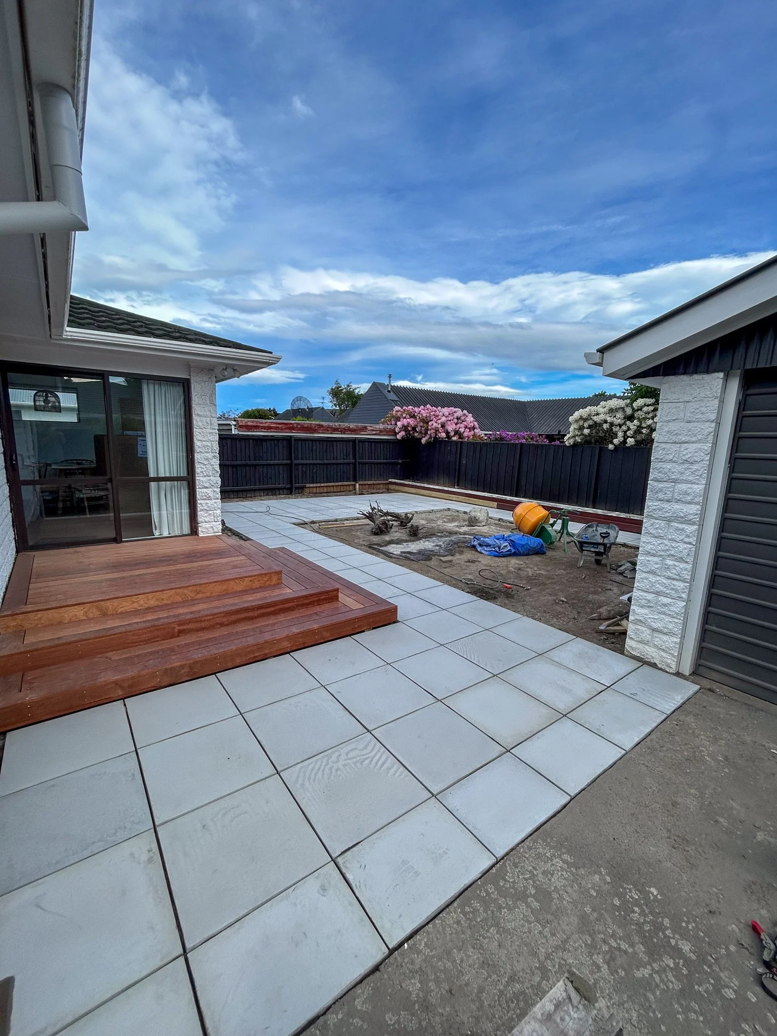 Backyard patio under construction with partially tiled floor, wooden steps, and construction tools and materials scattered around.