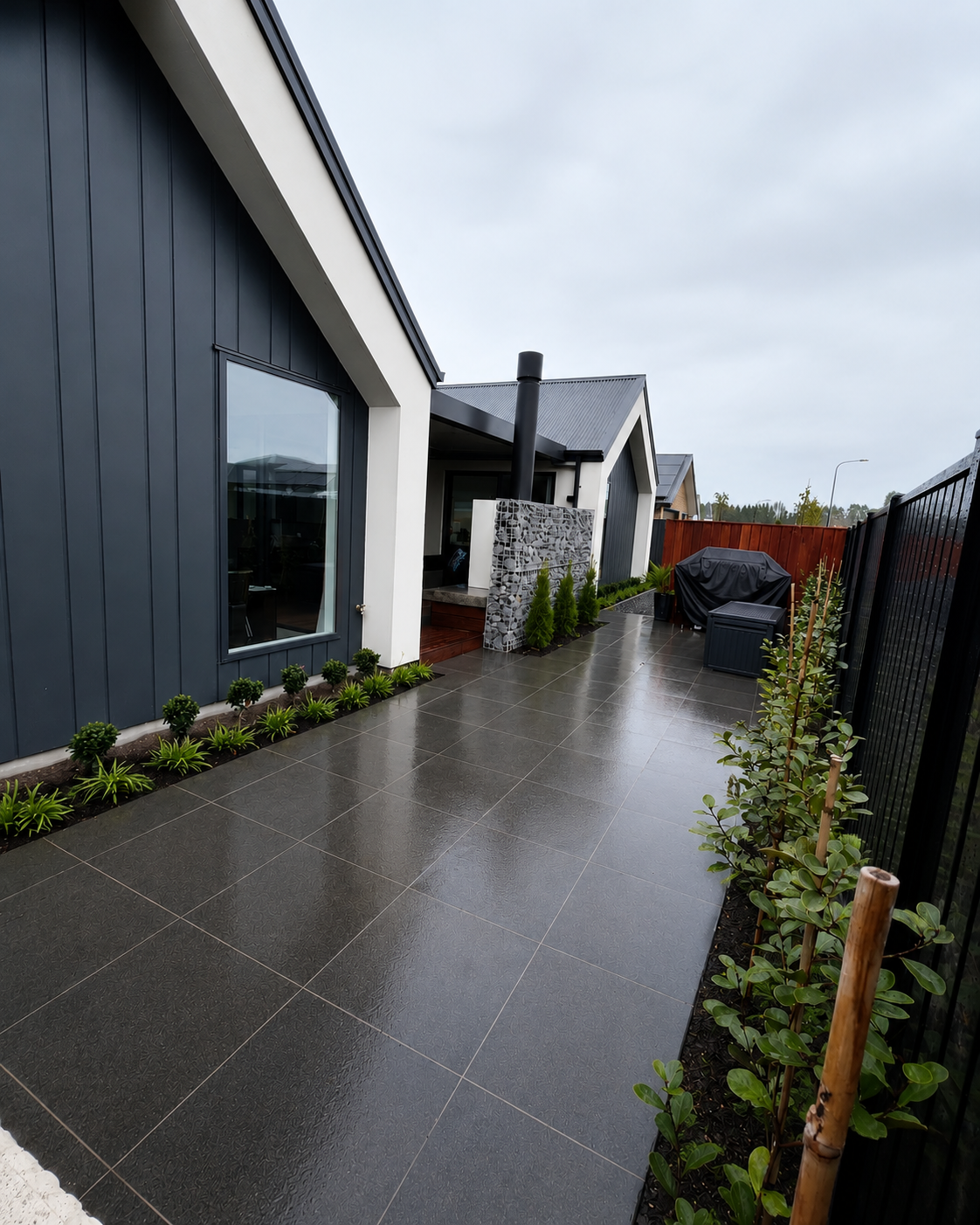 Modern backyard with dark tiled patio, small plants along the house, and a garden area with a black fence and outdoor furniture covered with a black cover.
