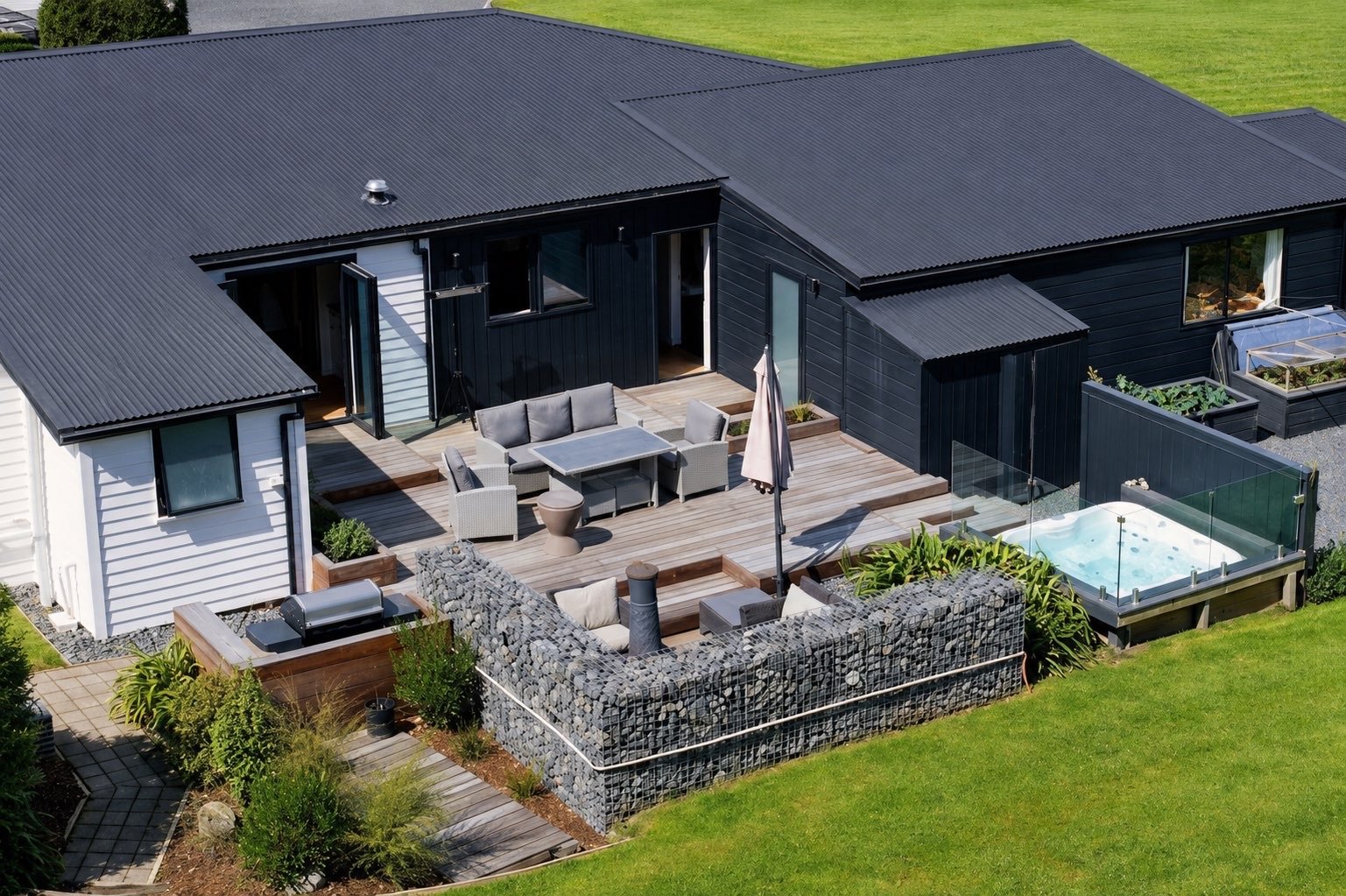 A modern house with a black metal roof and black exterior walls, featuring a spacious outdoor deck with seating, an umbrella, a hot tub, and surrounding greenery.