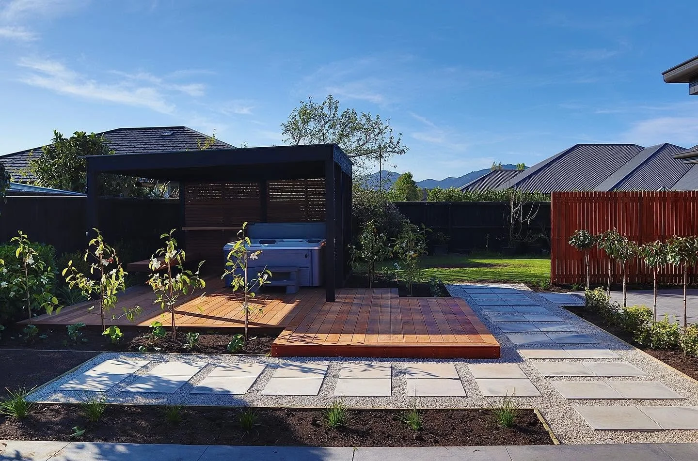 Backyard with landscaped garden, gray stone pathway, wooden deck, small trees, and a privacy fence under clear blue sky.