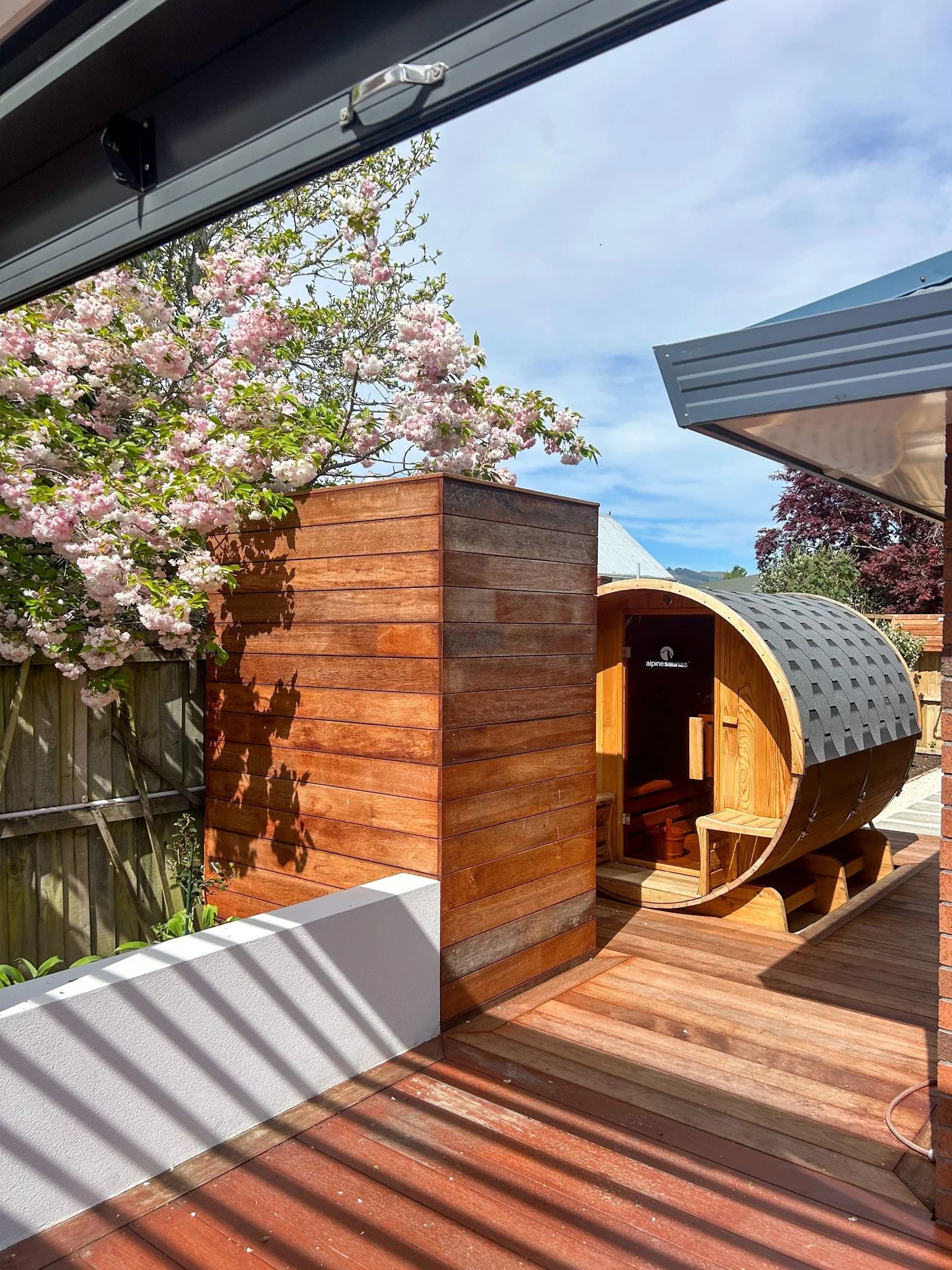 A small wooden outdoor sauna shaped like a barrel with a sign reading 'alpinesaunas' on the door, located on a wooden deck with a fence and blooming pink cherry blossom tree in the background, under a blue sky with some clouds.