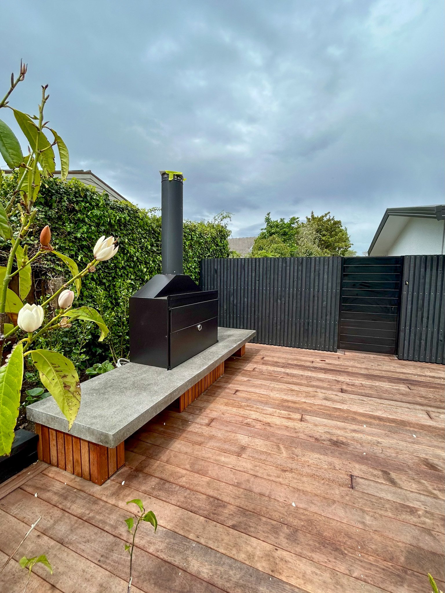 View of a backyard patio with wooden decking, black privacy fence, and lush greenery including a flowering plant with white blooms on the left side. The sky is cloudy.